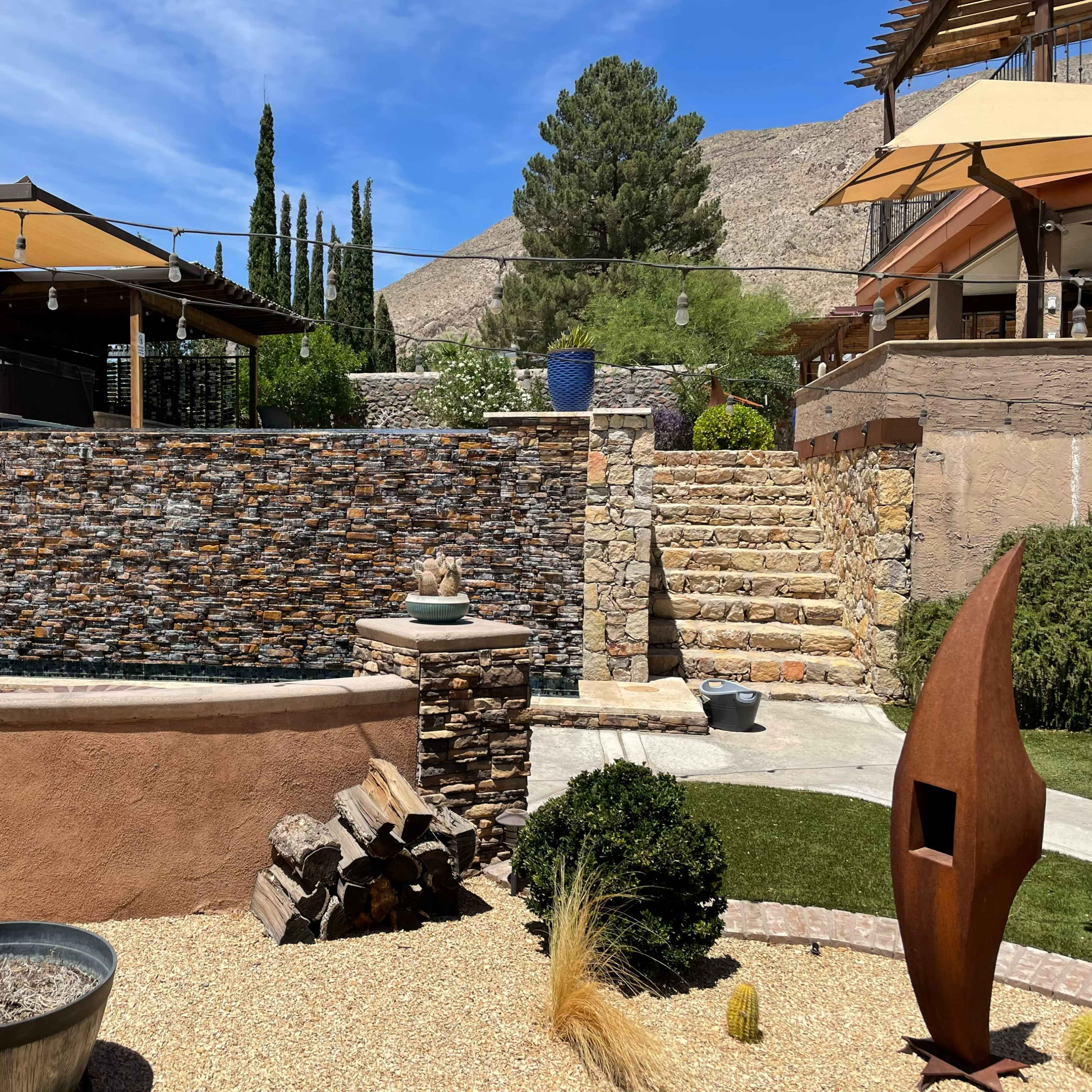 A landscaped outdoor area with a stone wall fountain, steps leading up to a structure, and various plants and decor.