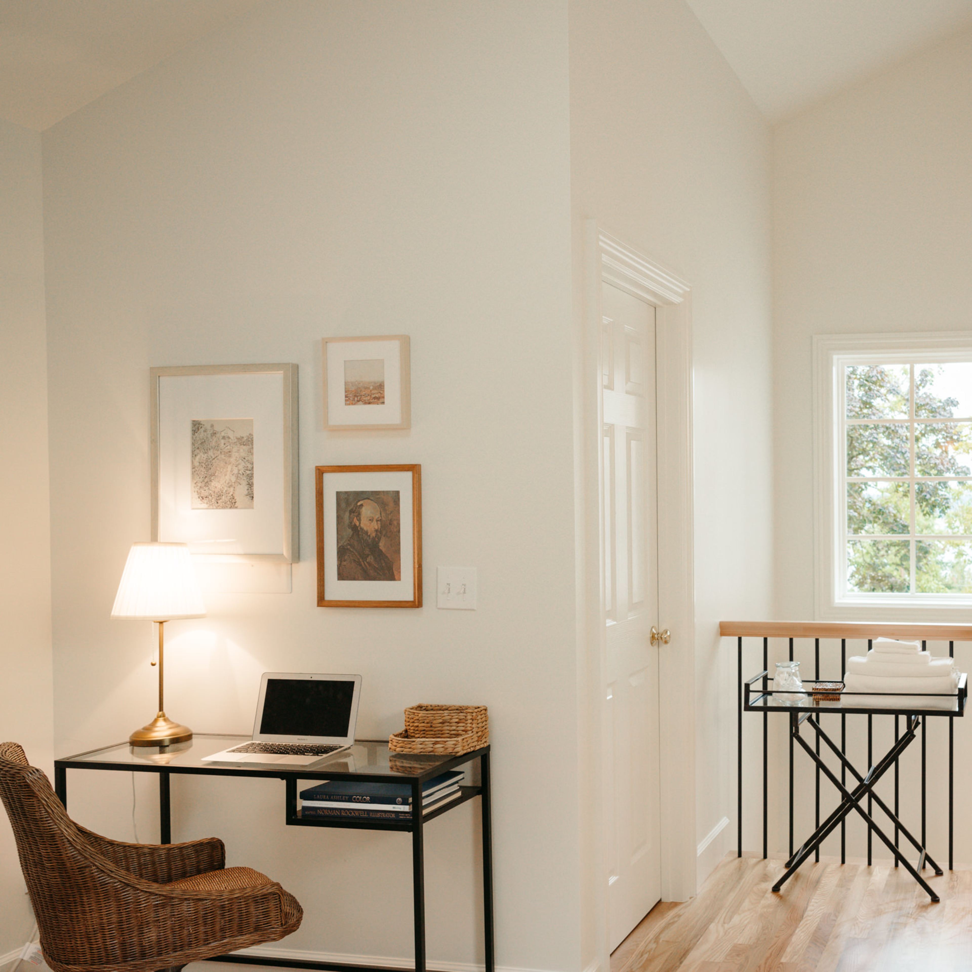 A well-lit corner of a minimalist interior features a desk with a laptop, a chair, and artwork on the wall, beside a window that provides a view of greenery outside.