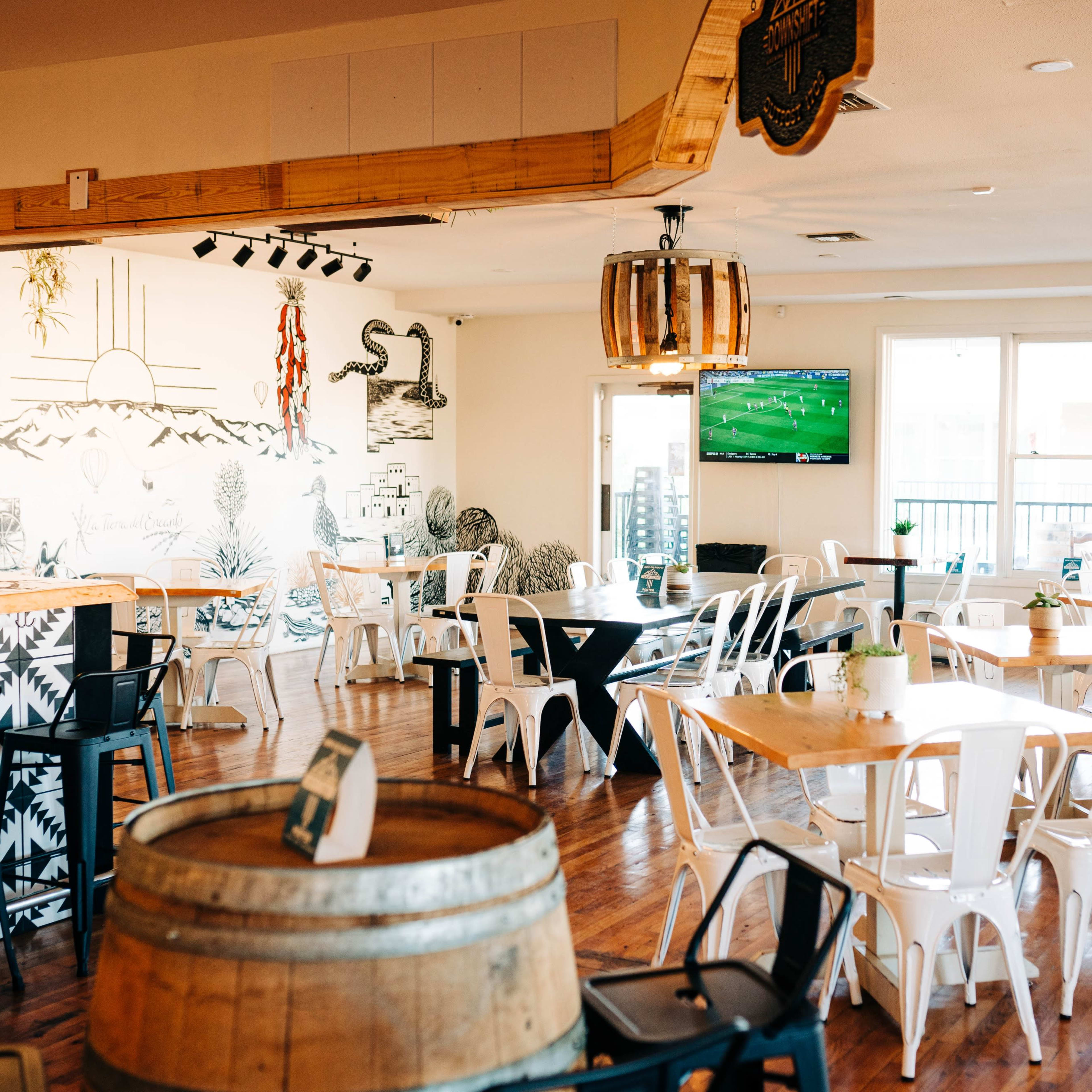 A spacious restaurant interior with wooden floors, tables set for dining, and a mural on the wall depicting mountains and a sunset.