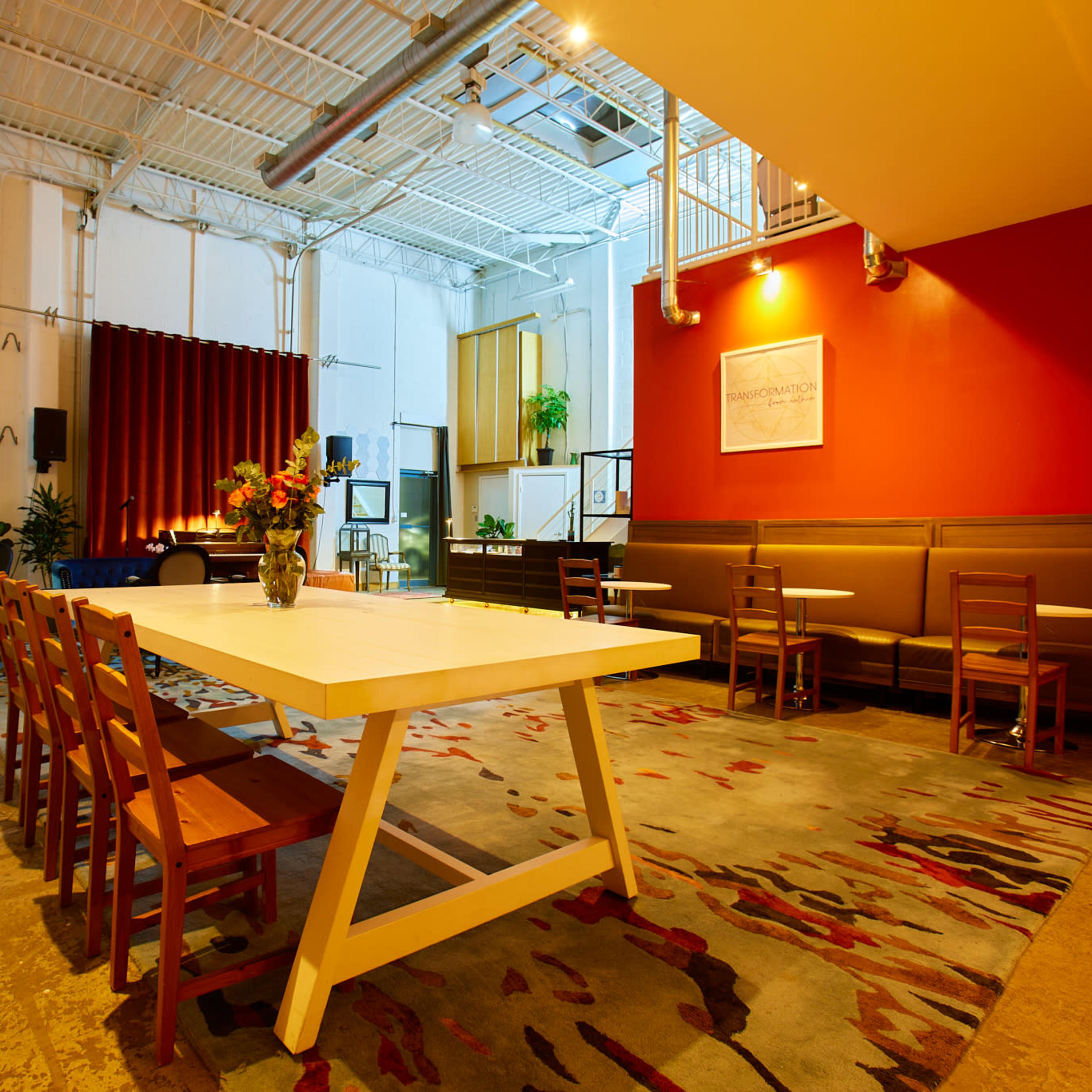 A well-lit, contemporary gathering space with a large table, several chairs, potted plants, and a stage area in the background featuring a red wall.