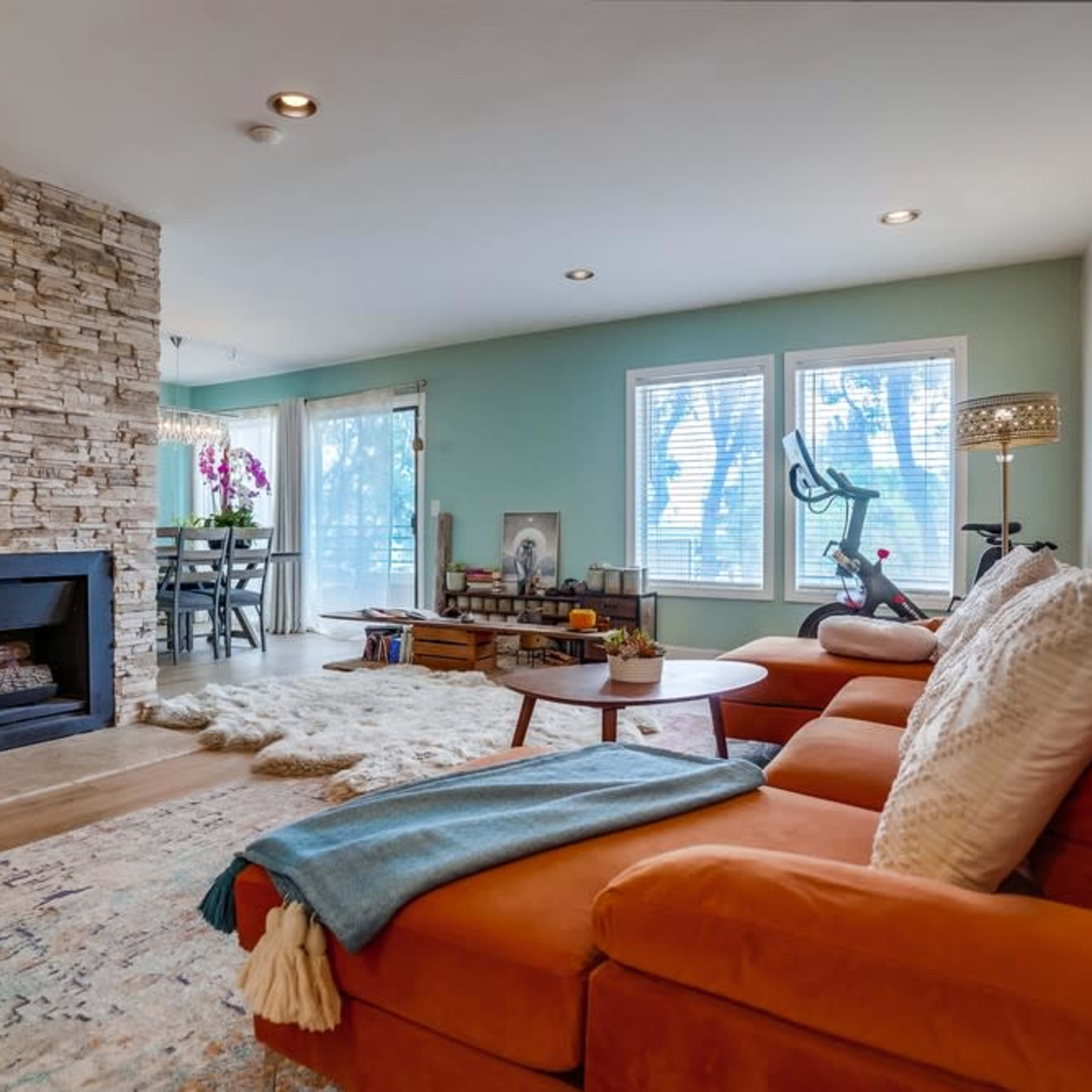 A cozy living room featuring an orange sectional couch, a stone fireplace, and large windows with a view of greenery outside.