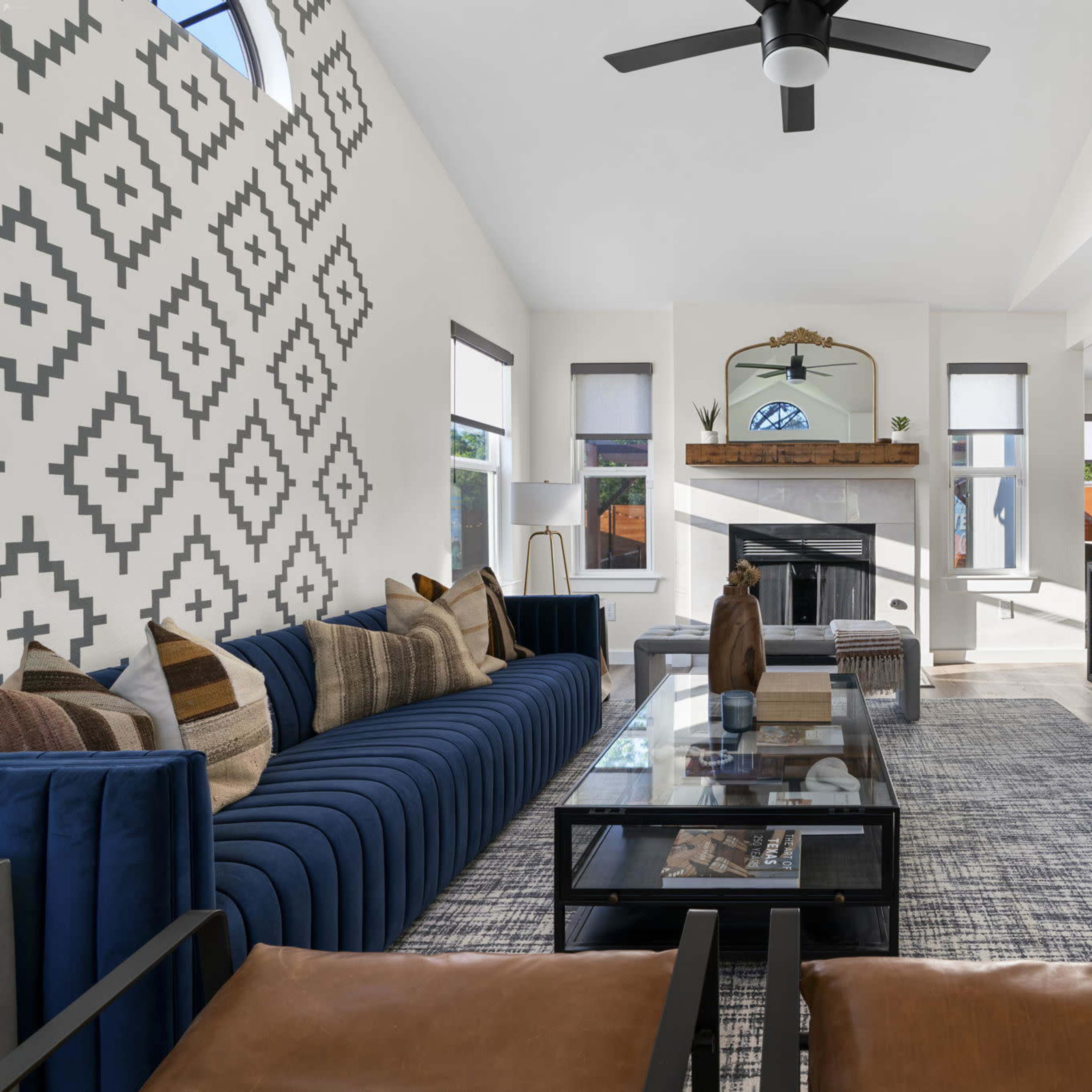 A modern living room features a blue sectional sofa, a glass coffee table, and a mounted television above a fireplace, with patterned walls and large windows allowing natural light.