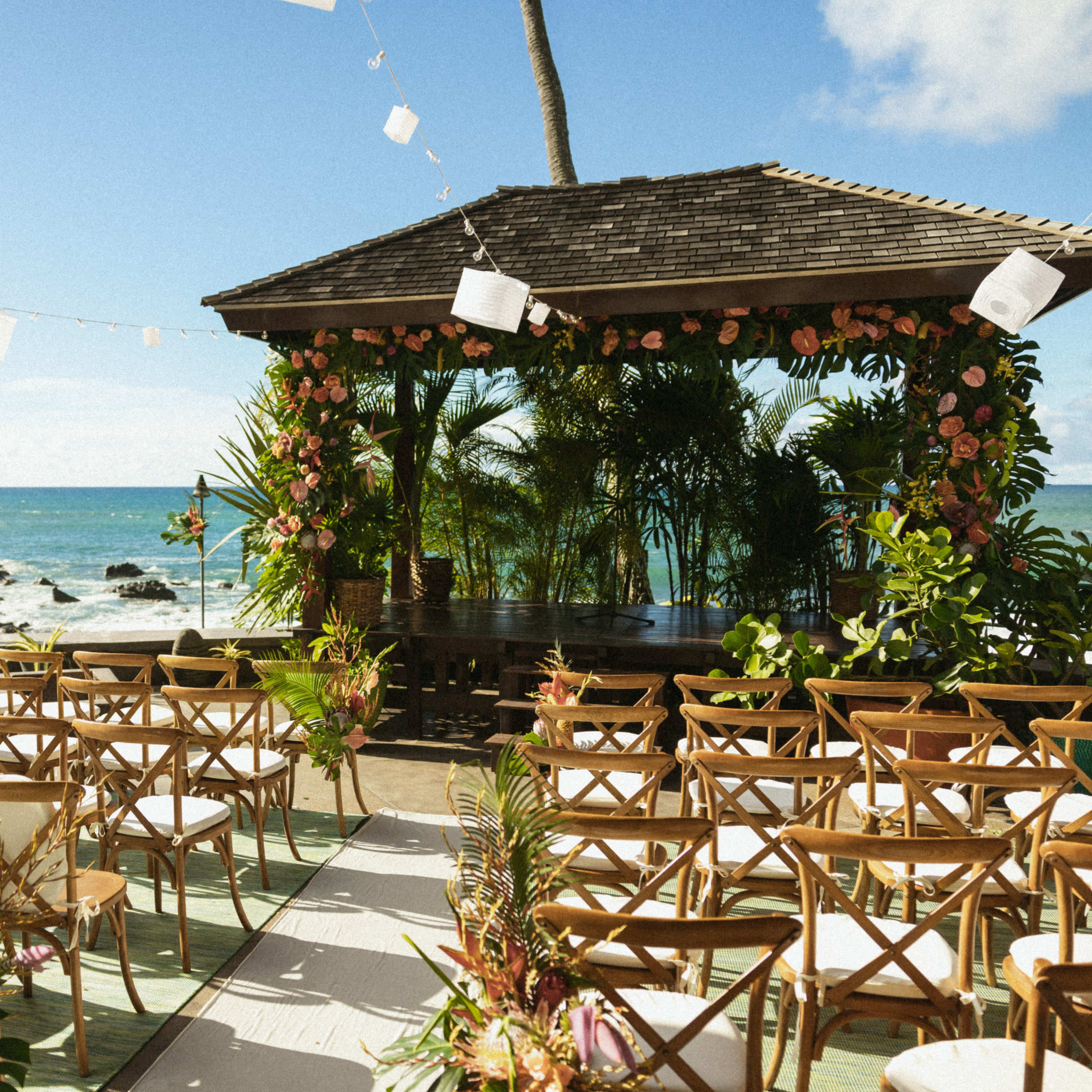 An outdoor wedding venue features rows of wooden chairs facing a decorated pavilion with a view of the ocean.