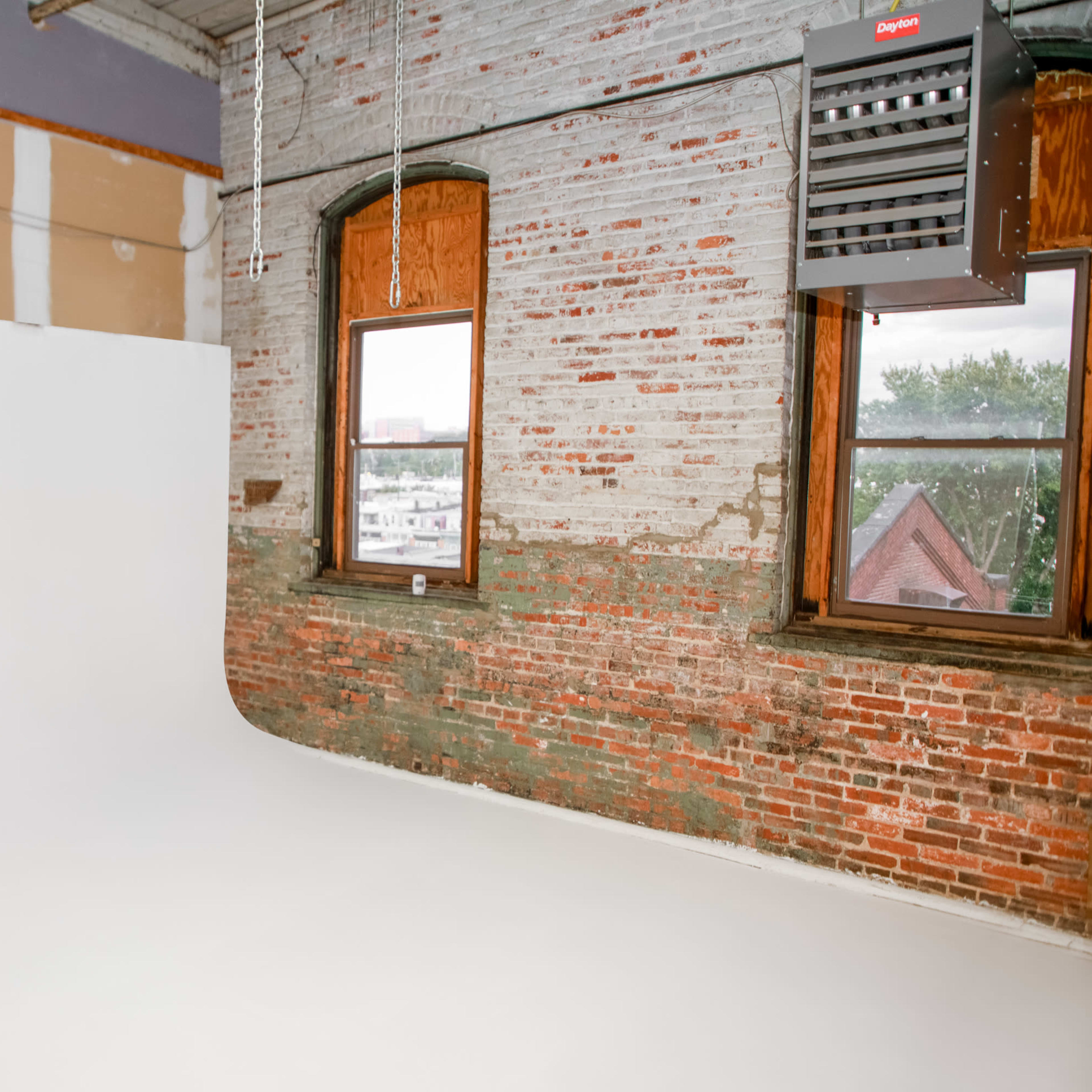 A large, empty photography studio features a seamless white backdrop and exposed brick walls with a window revealing a view outside.