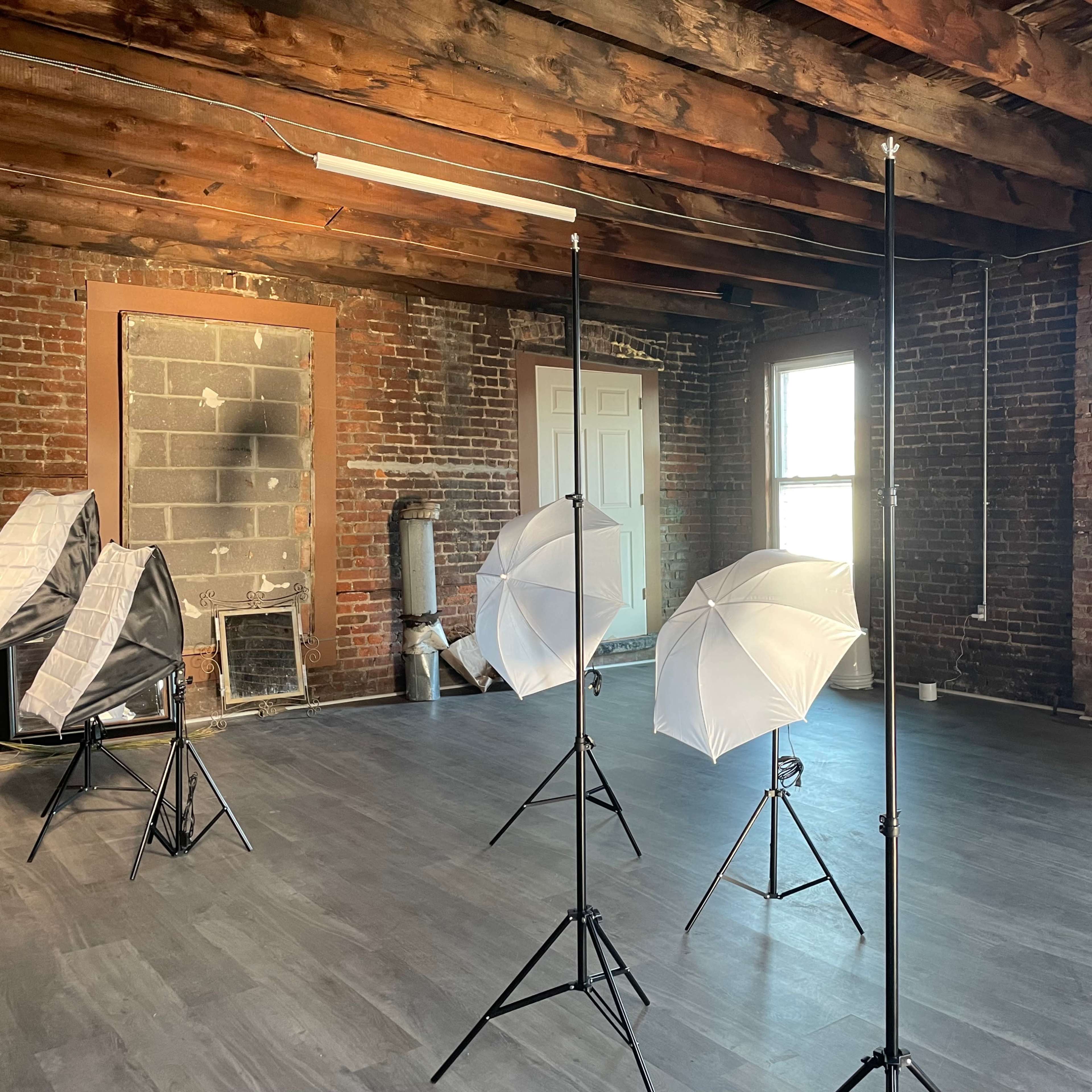 An empty room with exposed brick walls, wooden beams, and three photo lighting setups with white umbrellas.