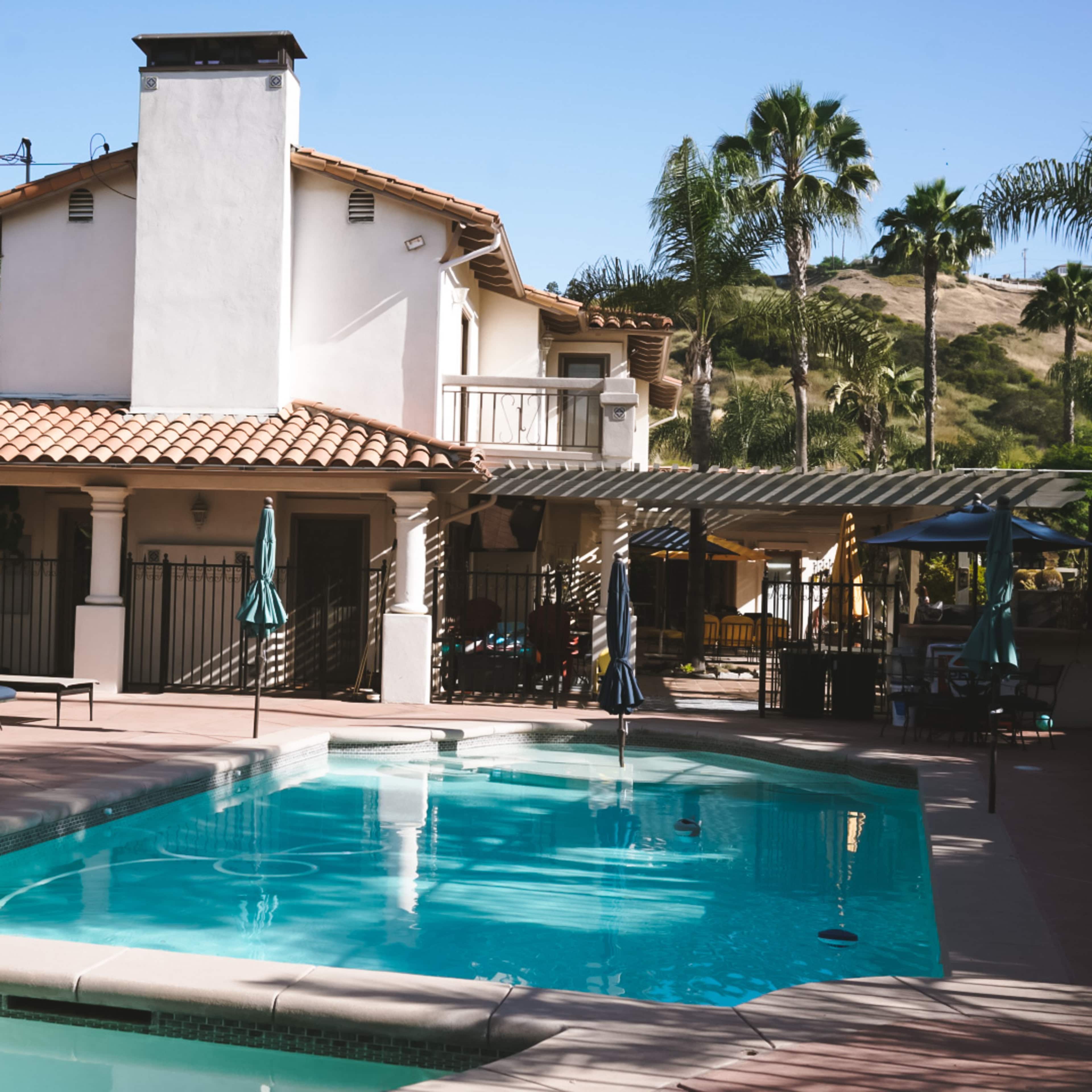 A two-story house with a tiled roof overlooks a pool surrounded by lounge chairs and palm trees.