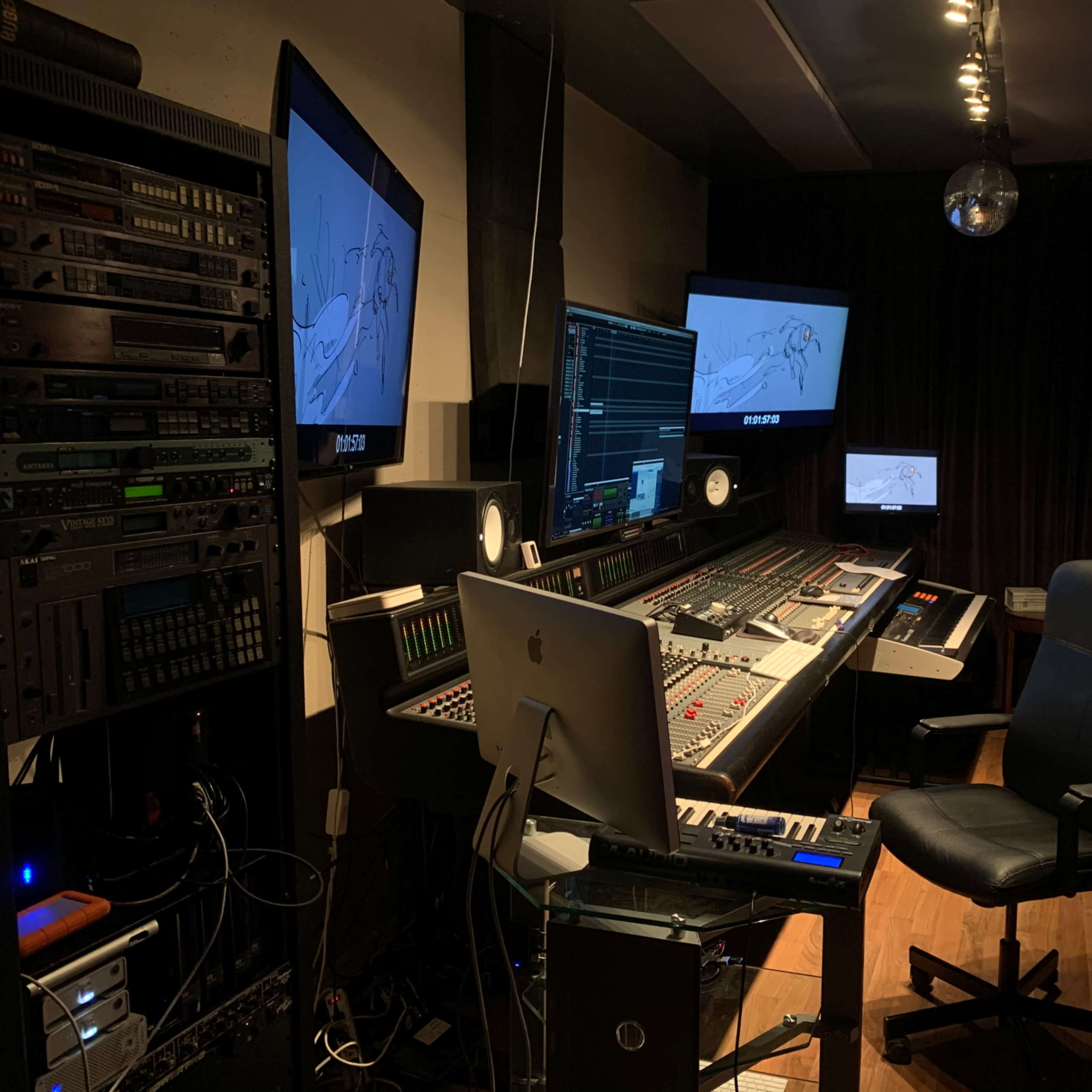 A music production studio equipped with a mixing console, two large monitors displaying audio waveforms, and various audio equipment on the walls.