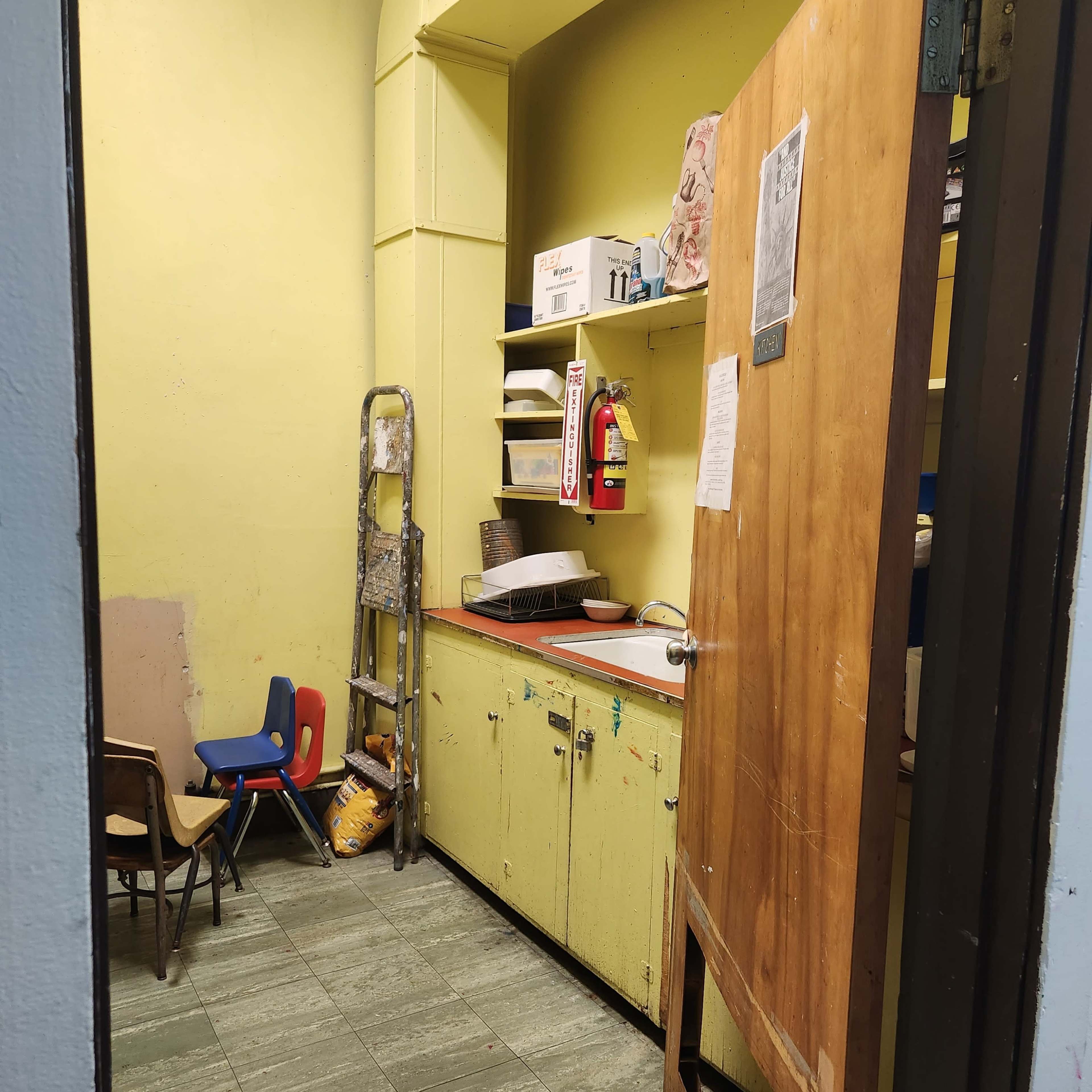 A small storage room with a wooden door ajar, empty shelves, a ladder, and children's chairs against the wall.