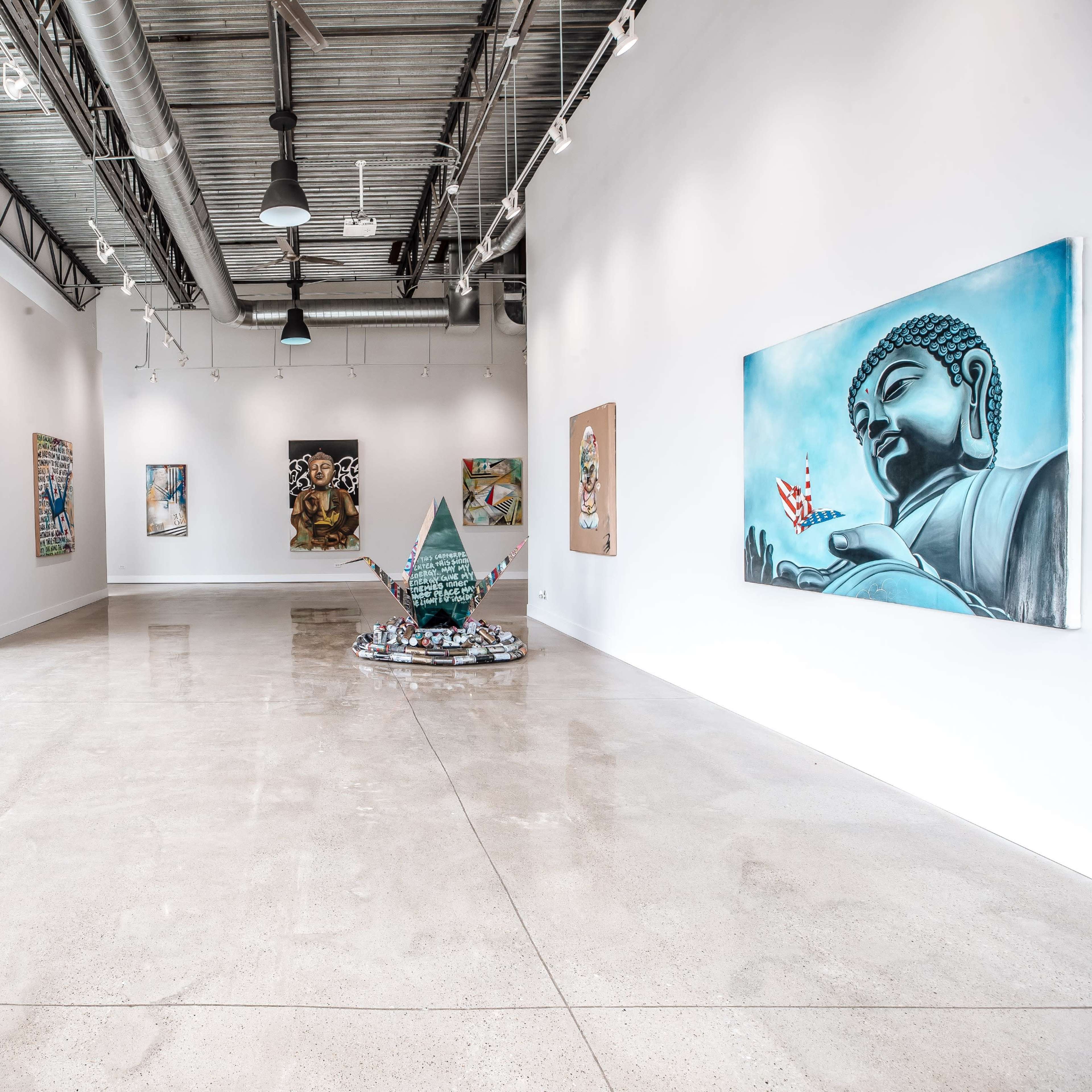 A spacious art gallery featuring various artworks, including a Buddha painting and a three-dimensional sculpture, with large windows allowing natural light to fill the room.