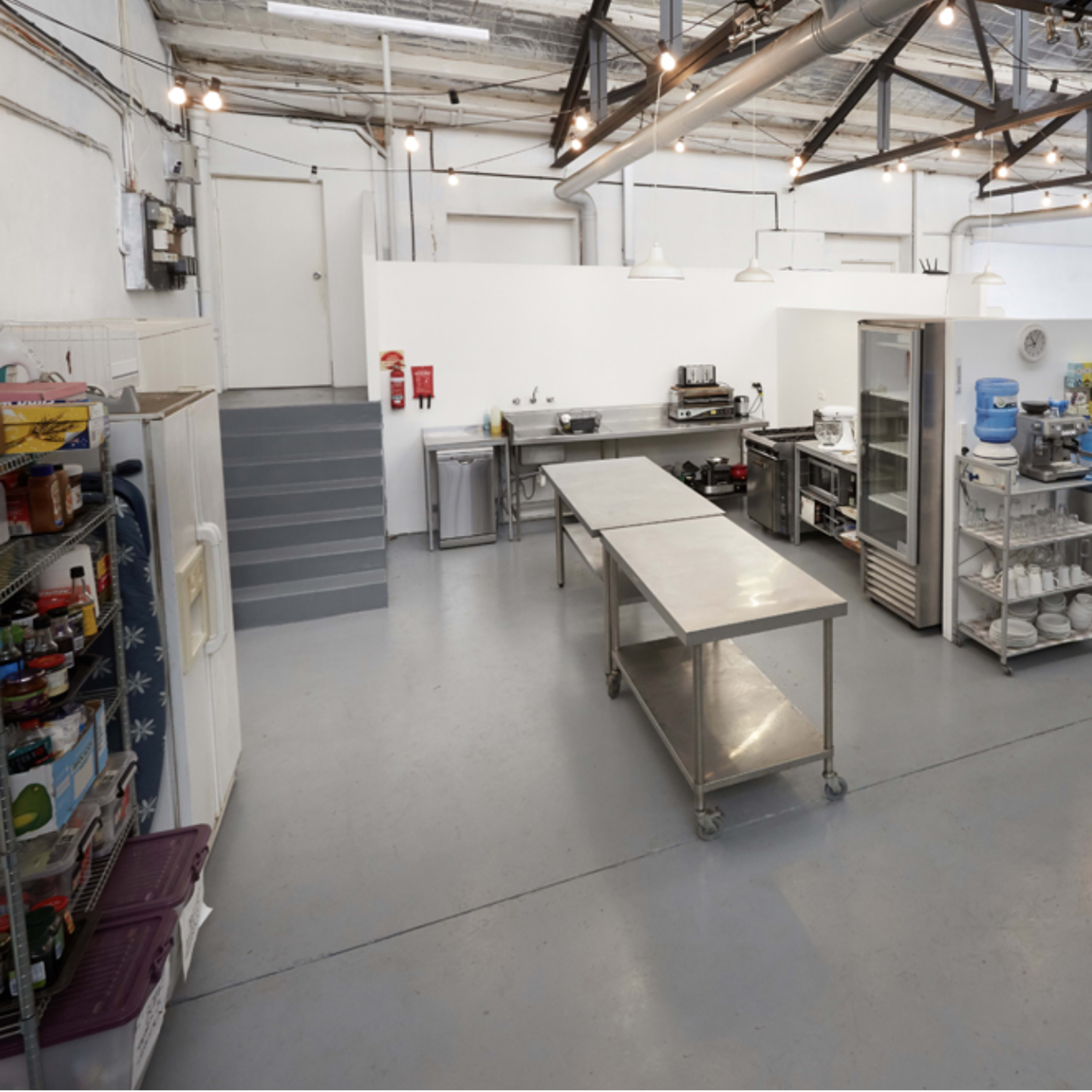 A spacious kitchen with stainless steel tables, appliances, and a refrigerator, featuring industrial lighting and an organized layout.