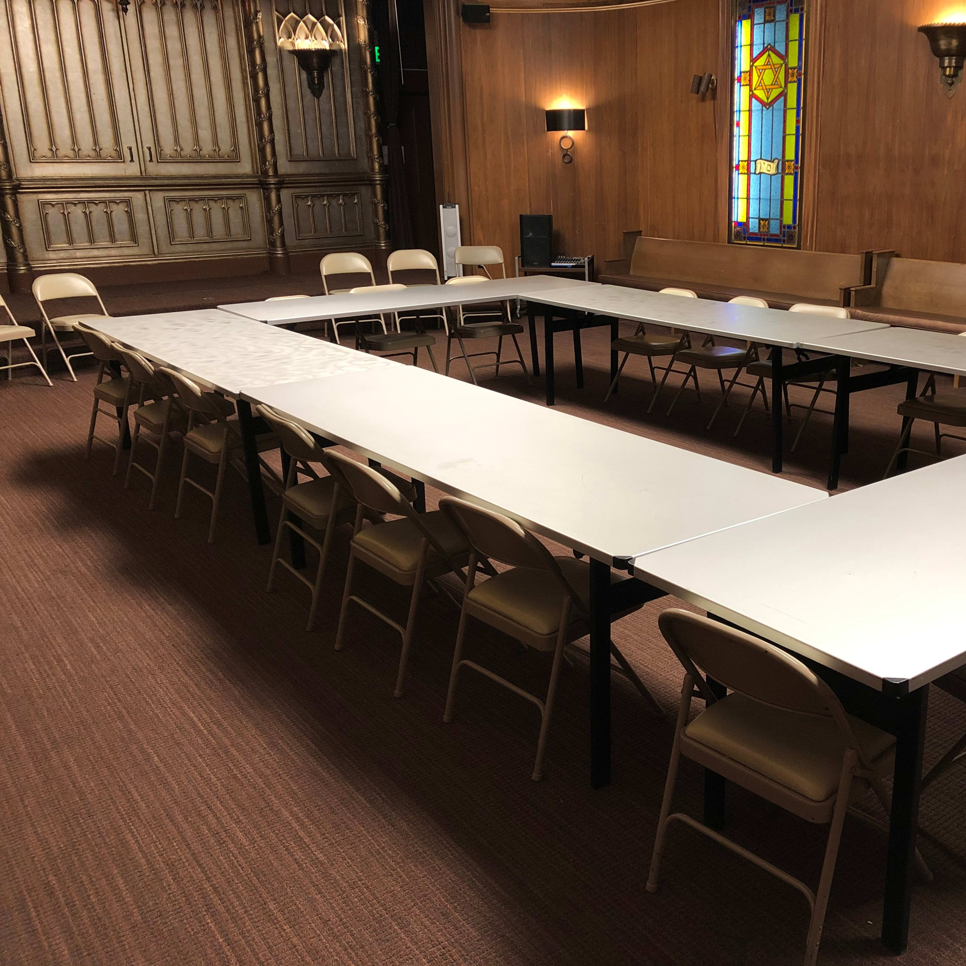 A large meeting room features an arrangement of tables in a U-shape, surrounded by folding chairs, with stained glass windows in the background.