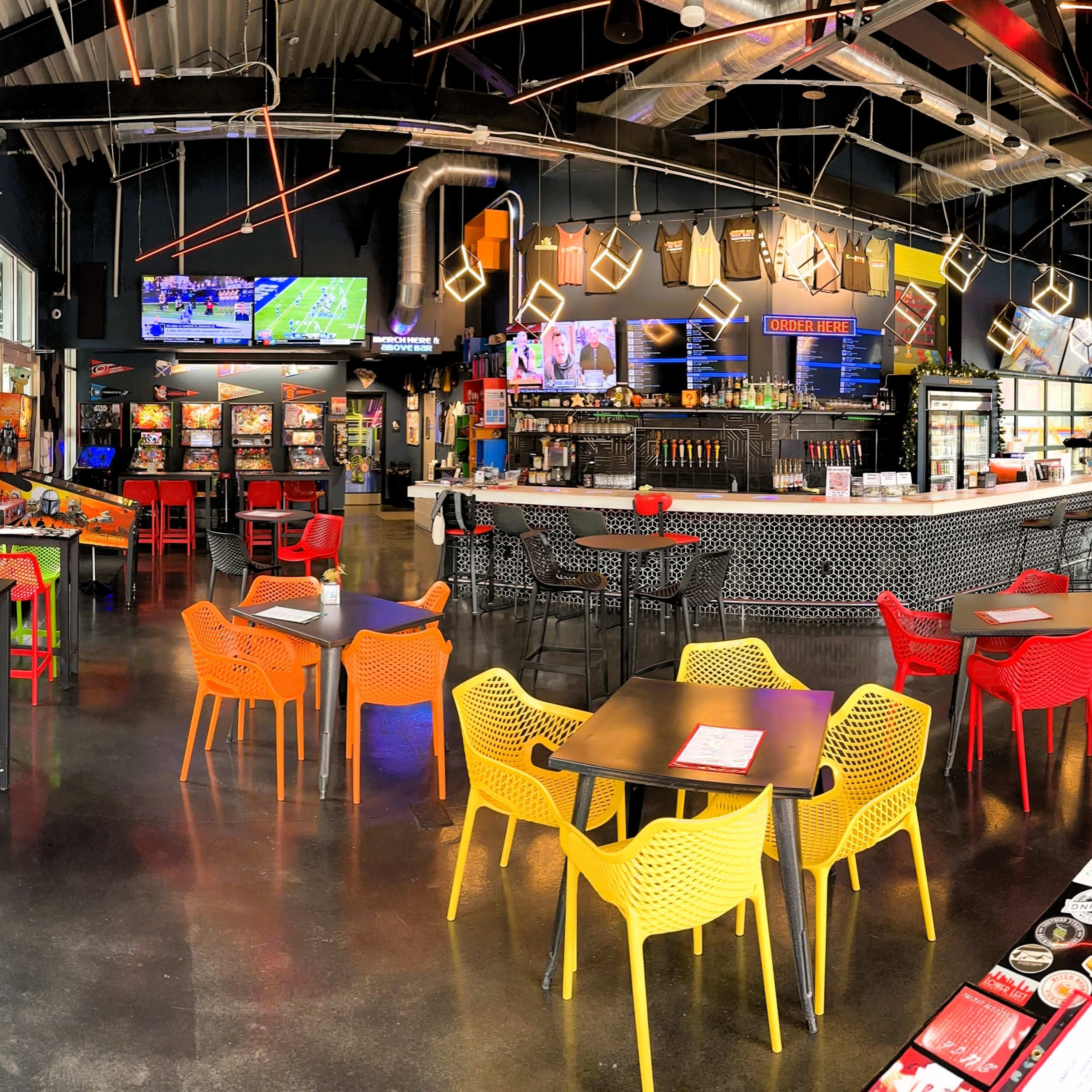 A modern bar and arcade with colorful chairs, a long counter, and various arcade machines and TVs displaying sports.