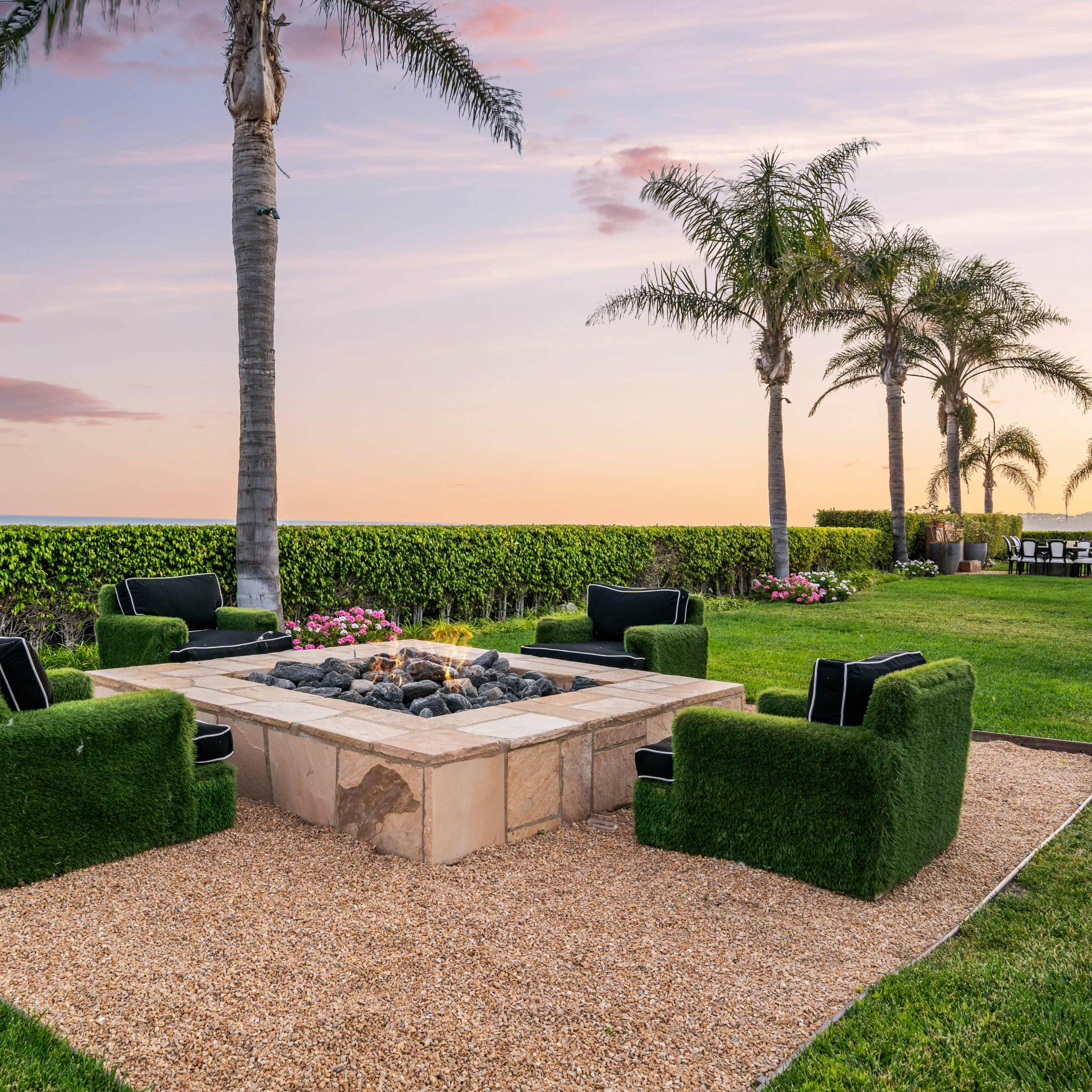 A circular stone fire pit surrounded by grass-covered seating is set in a landscaped area with palm trees and a view of the ocean at sunset.