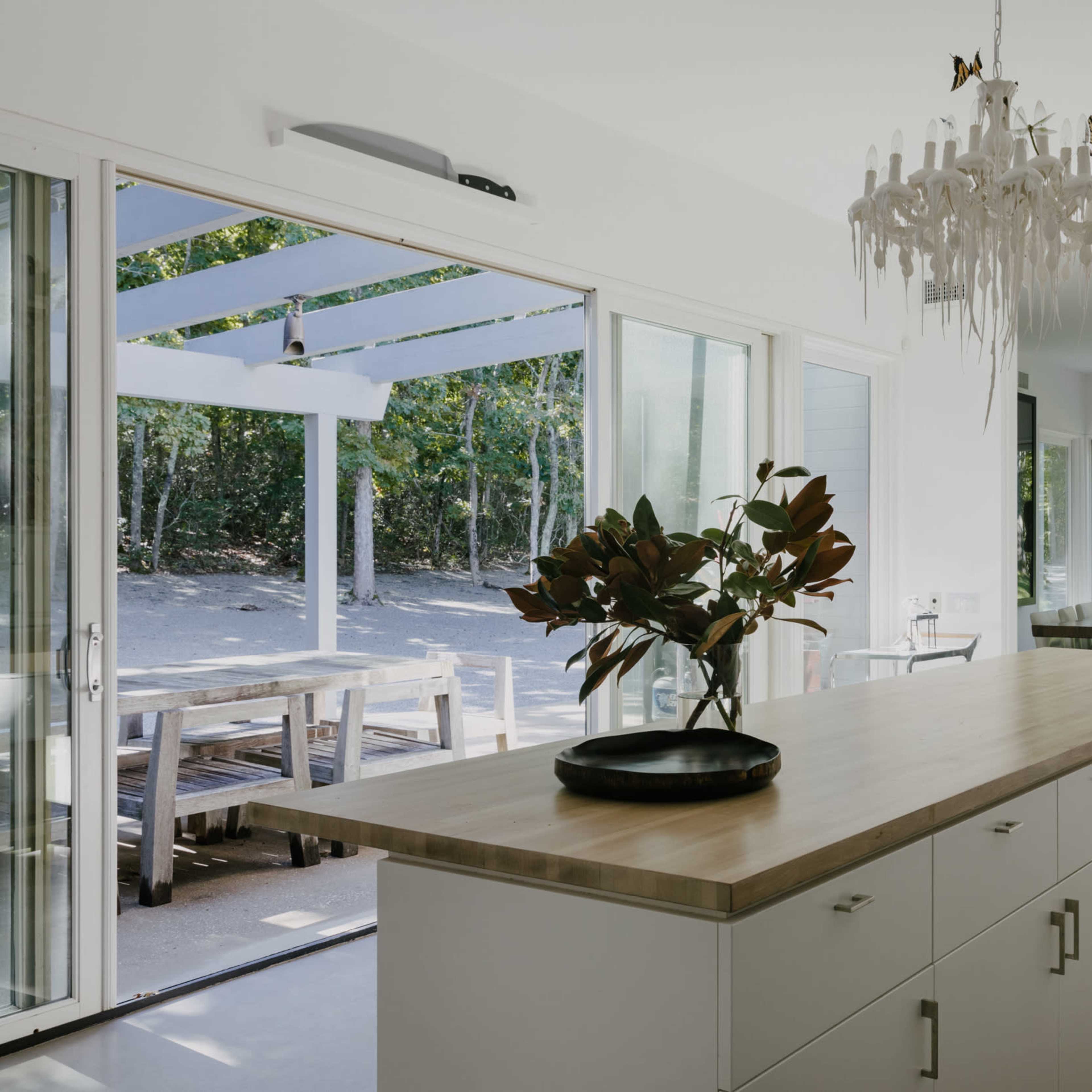 A modern kitchen features a wooden island, large windows overlooking an outdoor dining area, and a wall of open shelving displaying dishware.