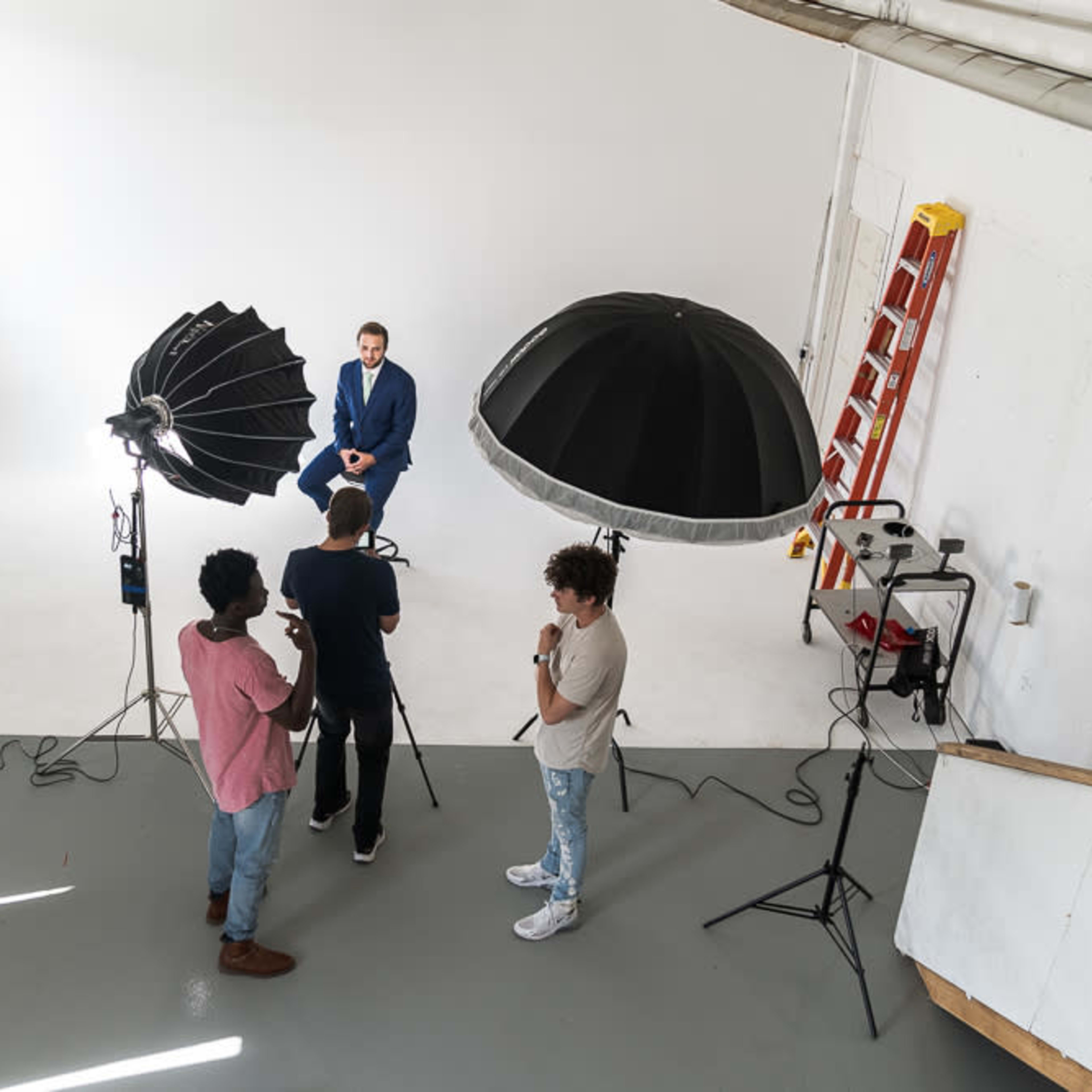A man in a blue suit is seated for a photoshoot while two other individuals set up equipment in a photography studio.