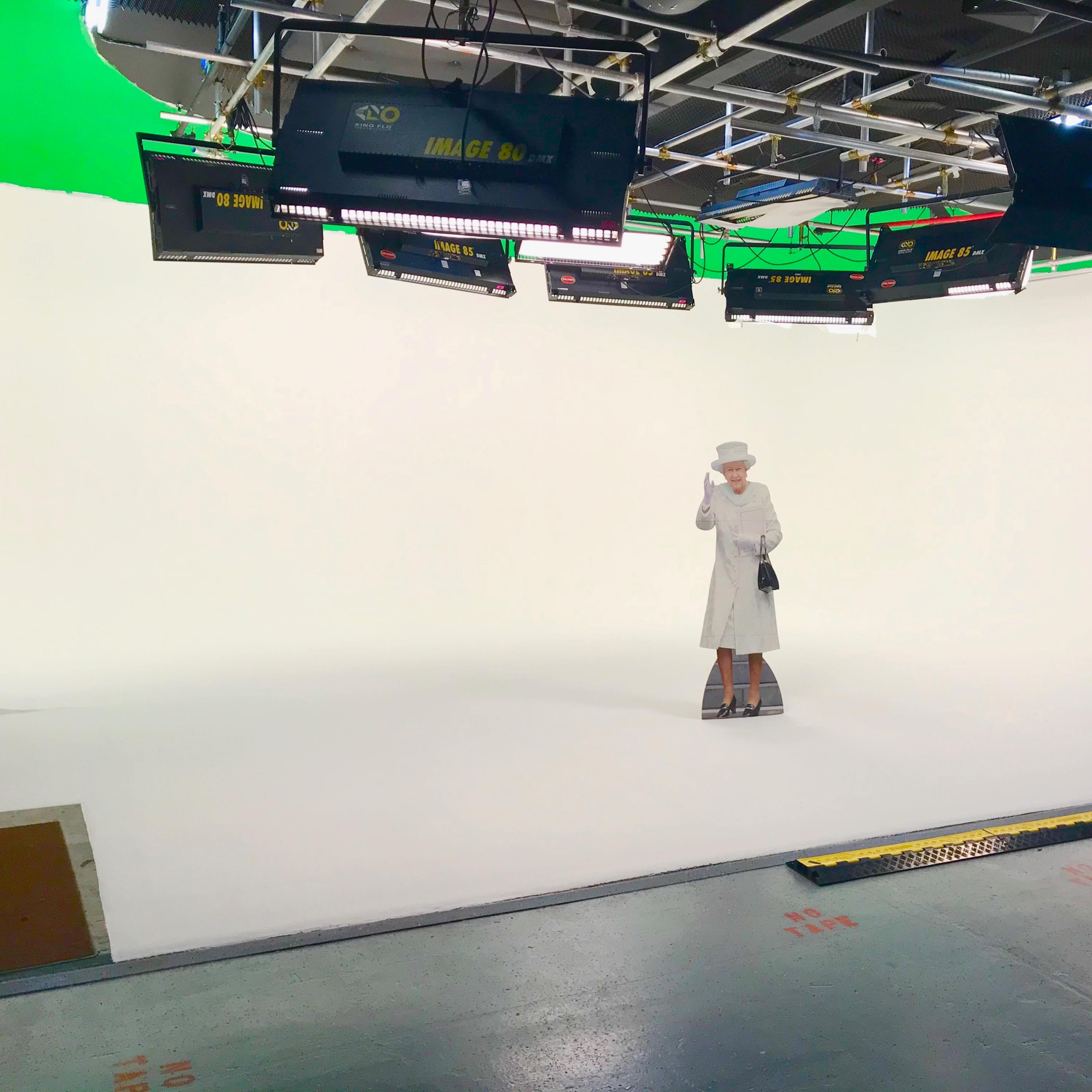 A lone figure dressed in a white outfit stands on a blank studio set, surrounded by bright lighting equipment and a green screen.