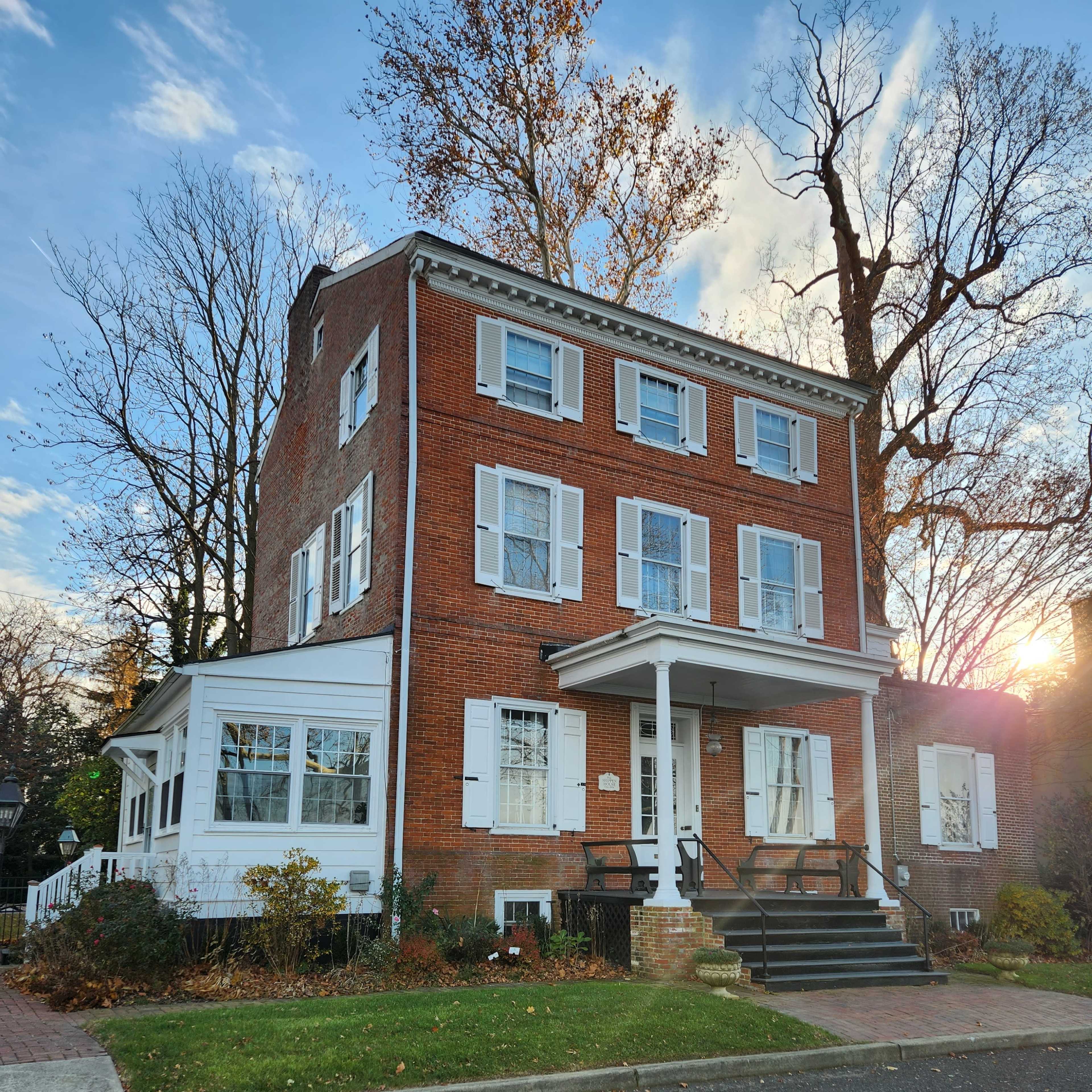 A three-story brick house with white shutters, a porch, and a large bay window, set on a tree-lined street during sunset.