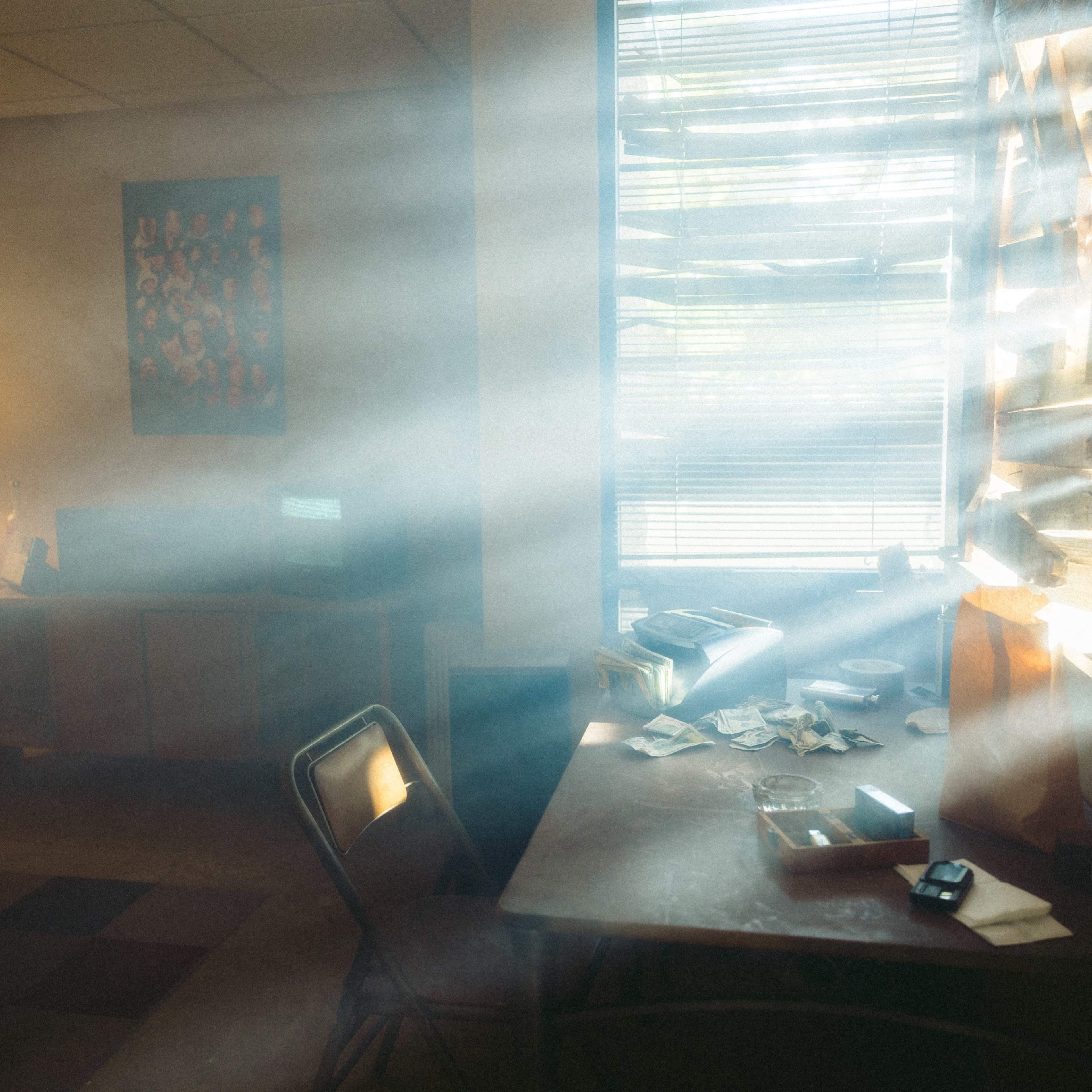 A dimly lit room with sunlight streaming through partially closed blinds, illuminating dust particles in the air and revealing a cluttered table and a couch.