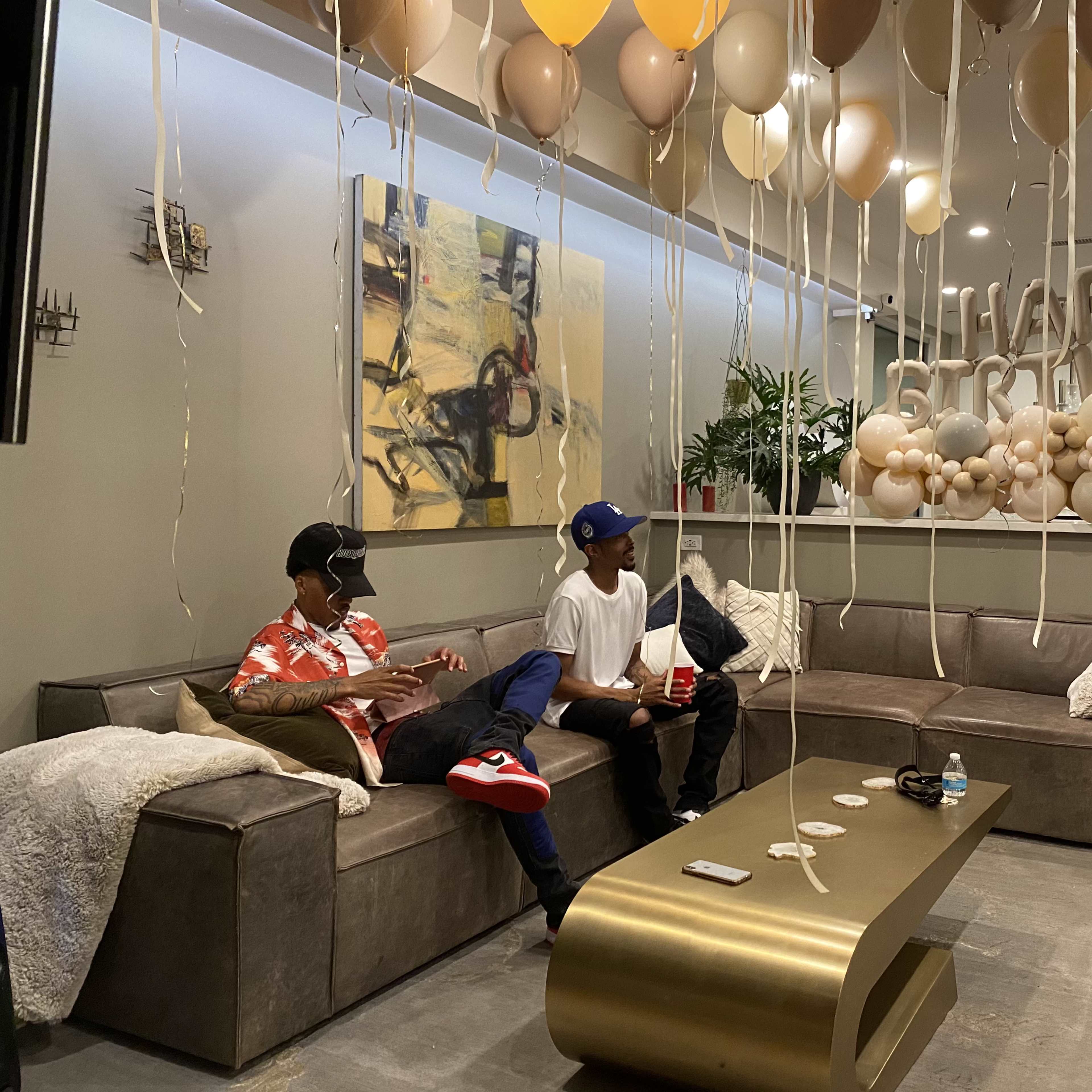 Two people sit on a couch in a decorated living room with balloons and a "Happy Birthday" display overhead.