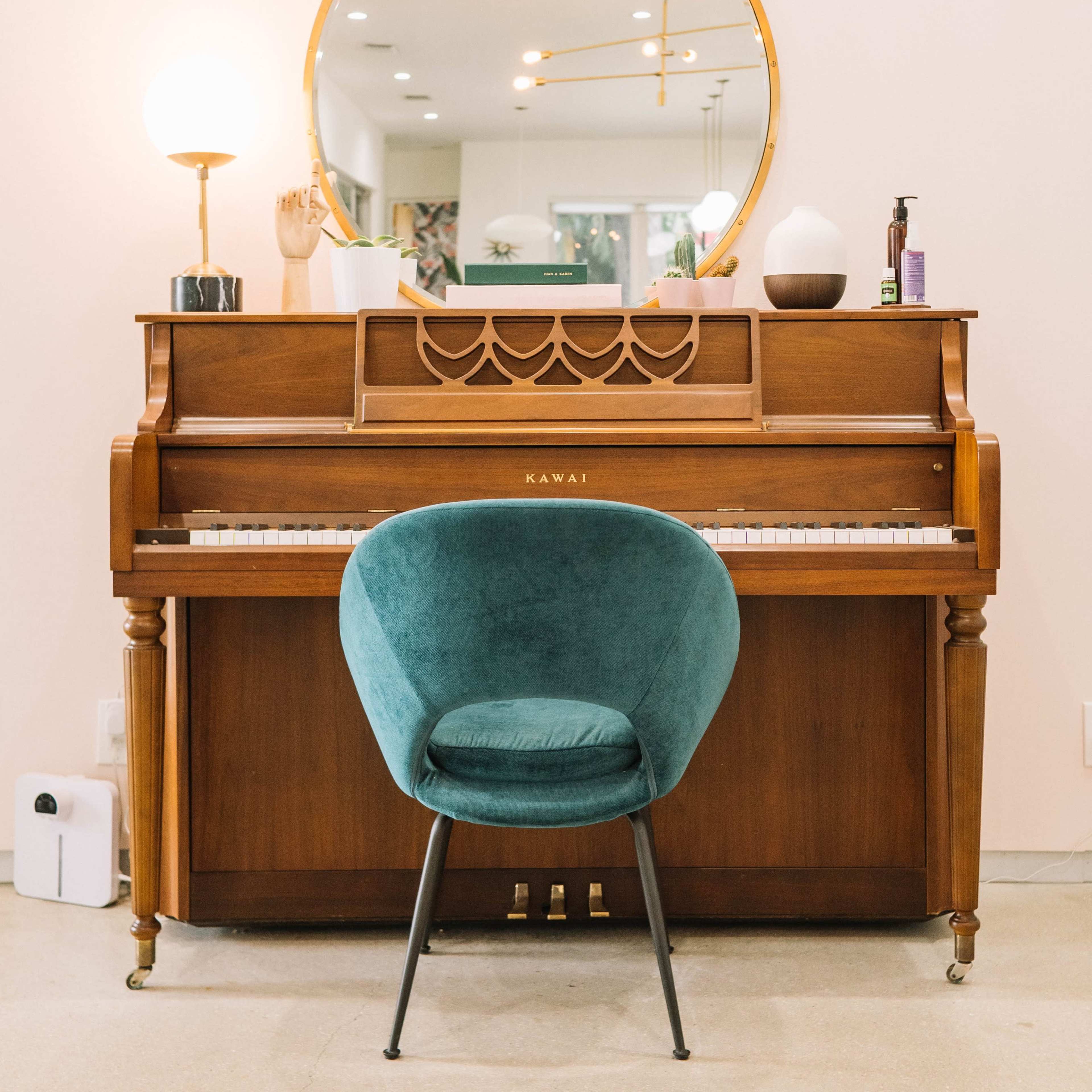 A wooden Kawai piano is positioned against a wall with a round mirror above it and a teal chair in front.
