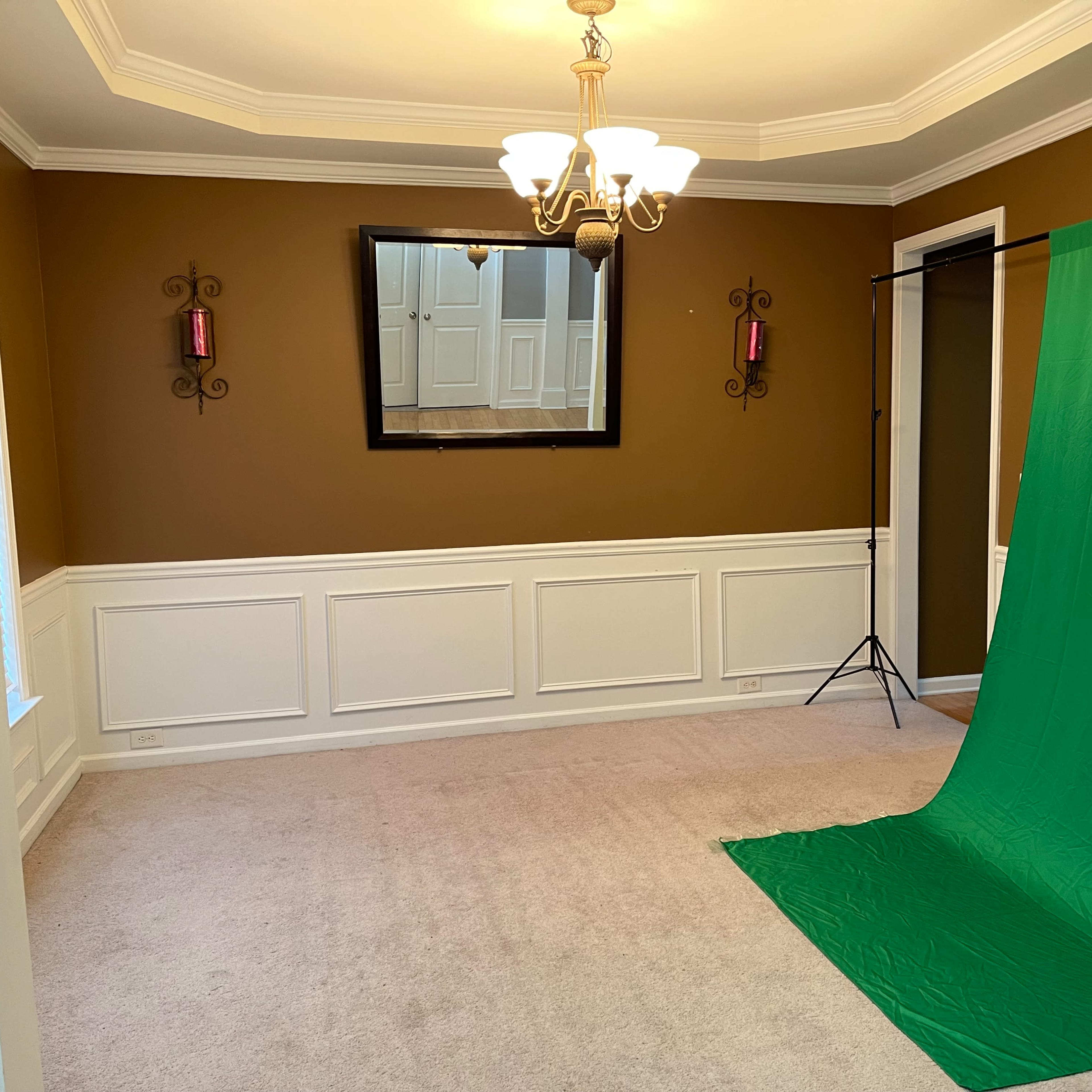 An empty room with brown walls, a chandelier, a large mirror, and a green backdrop set up in one corner.