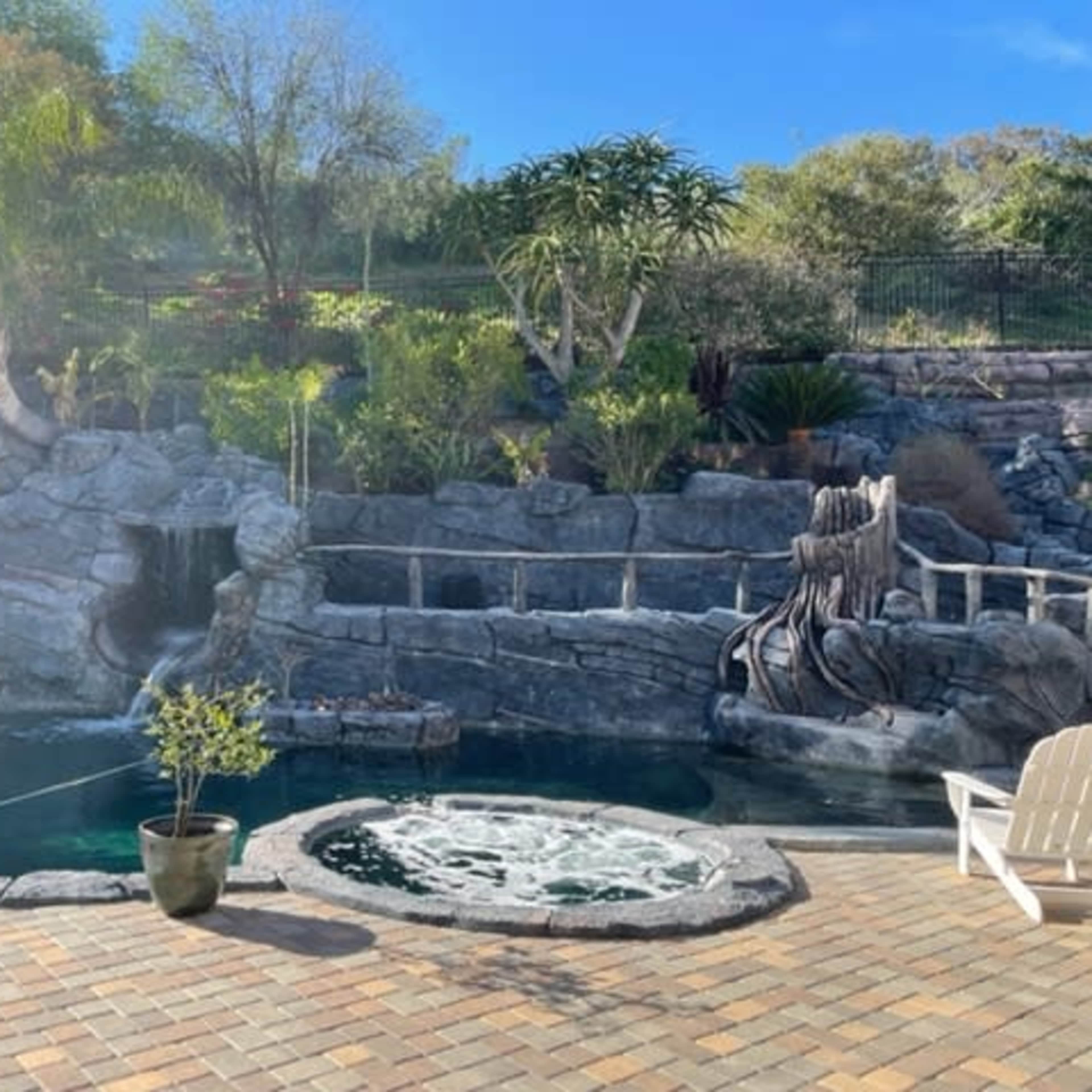 A landscaped backyard with a rock-themed swimming pool, a waterfall feature, and a hot tub surrounded by greenery.