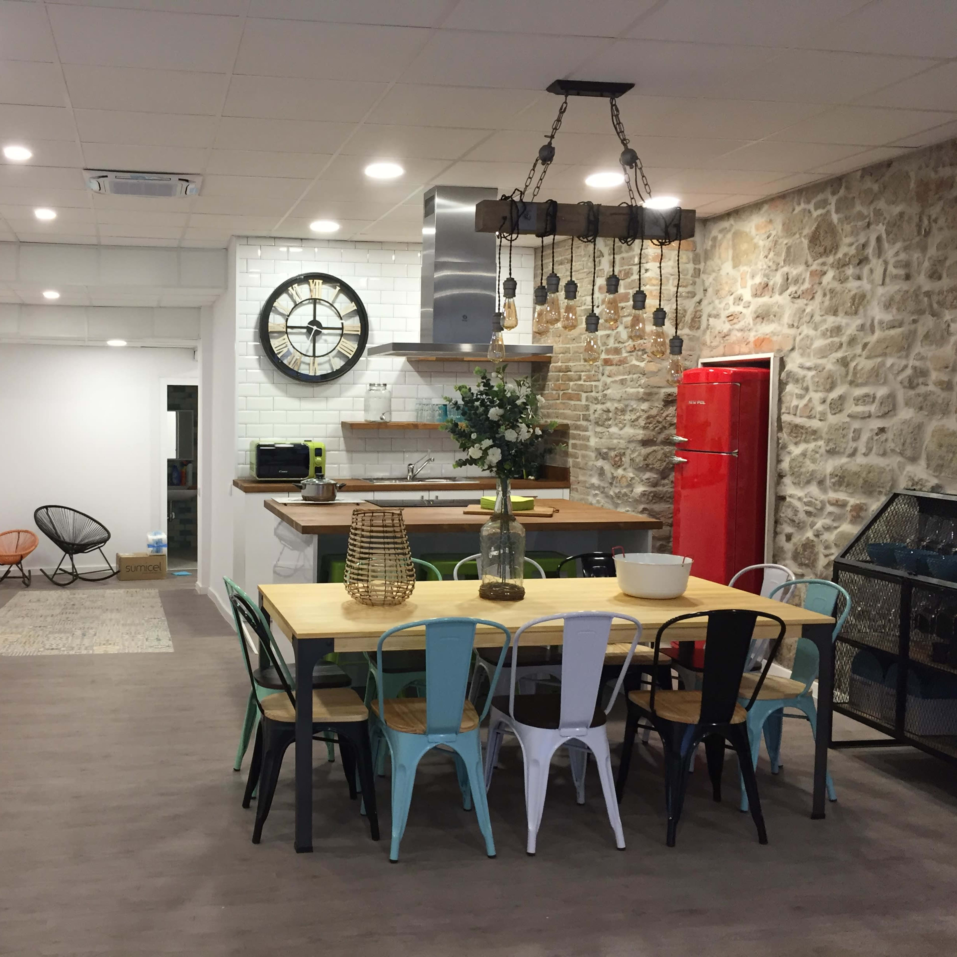 A modern kitchen and dining area features a large table with multicolored chairs, a red refrigerator, and a stone wall backdrop.