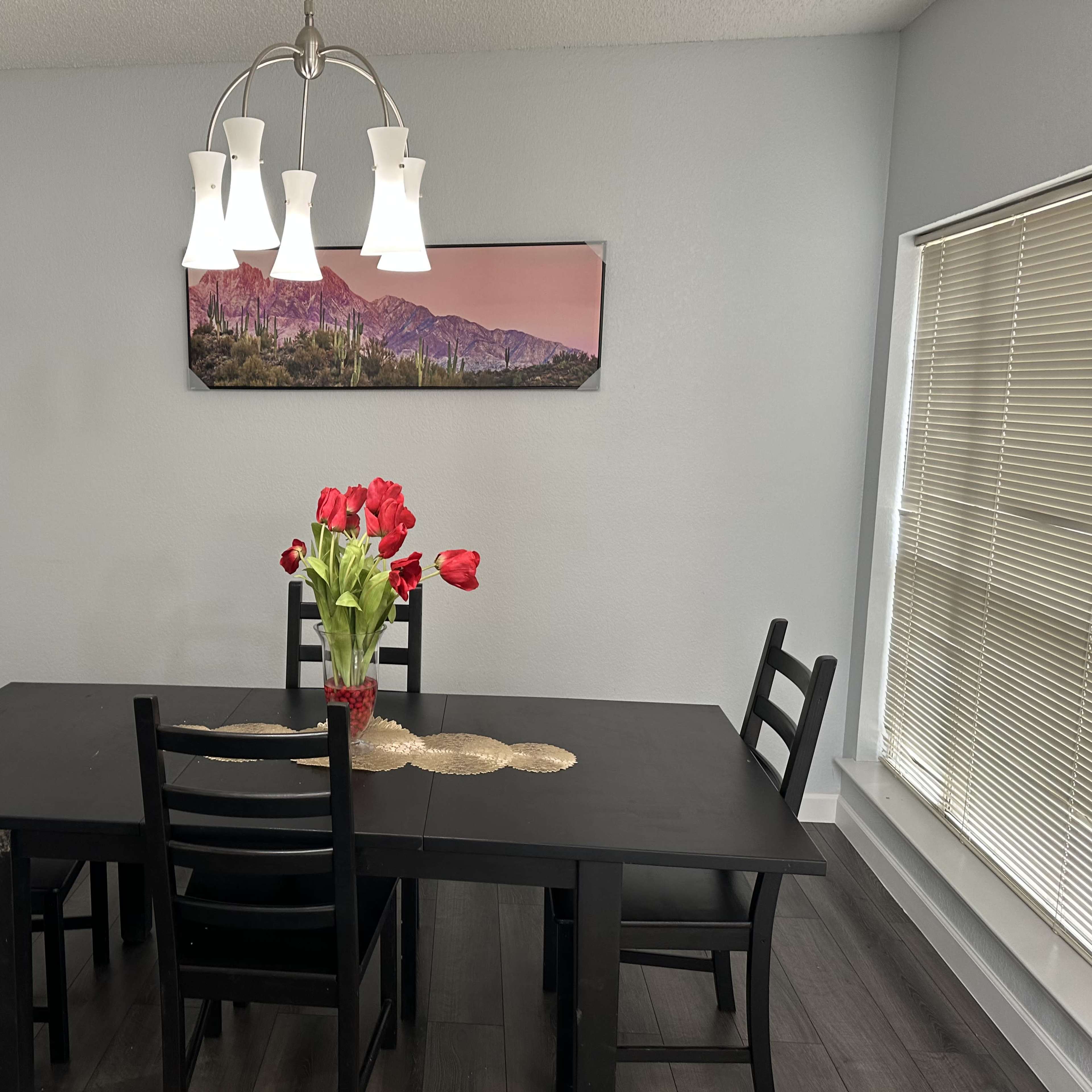 A dining area features a black table surrounded by chairs, a vase of red flowers, and a pendant light above, with a wall art piece depicting mountains.