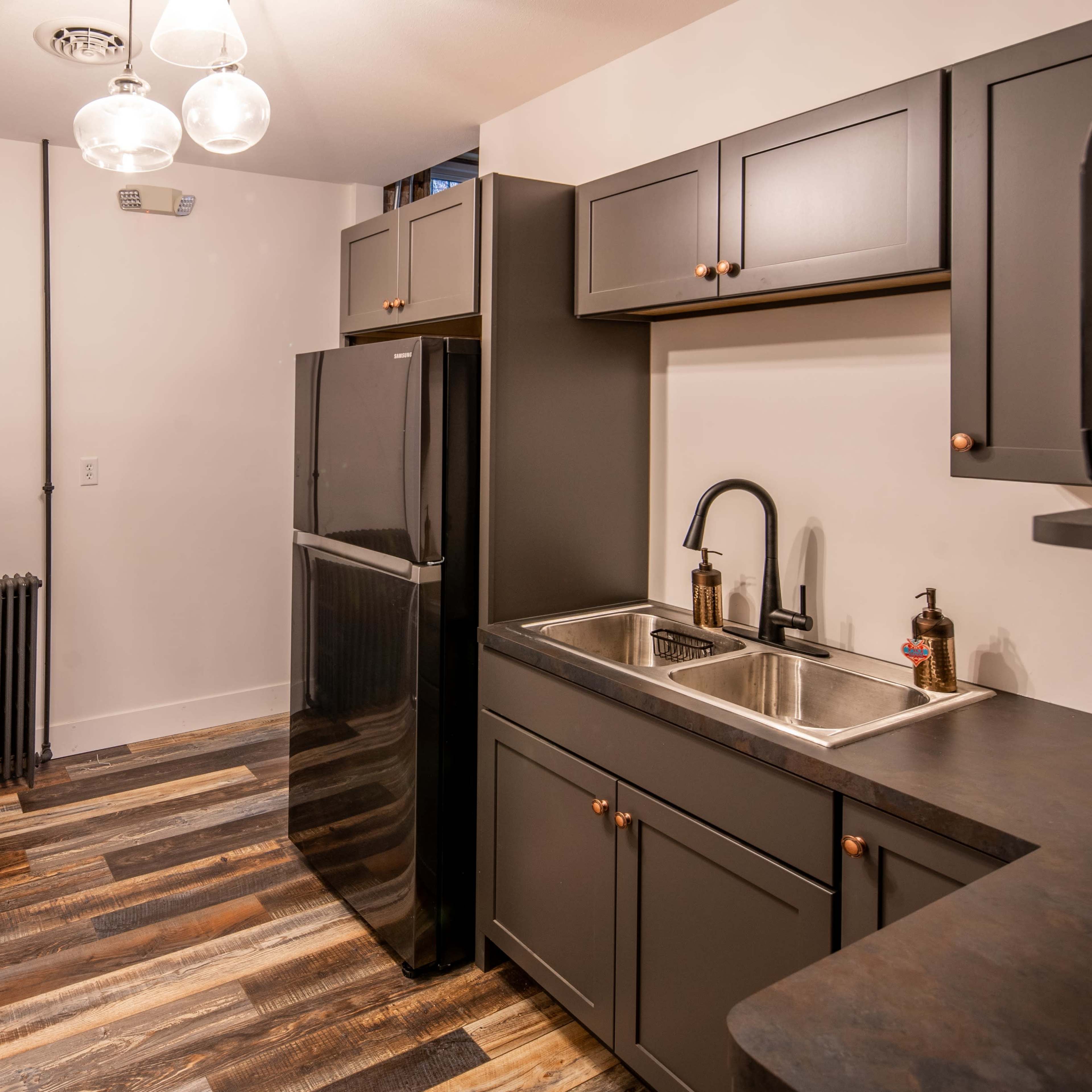 A modern kitchen features a black refrigerator, stainless steel sink, and dark cabinetry with hardwood floors.