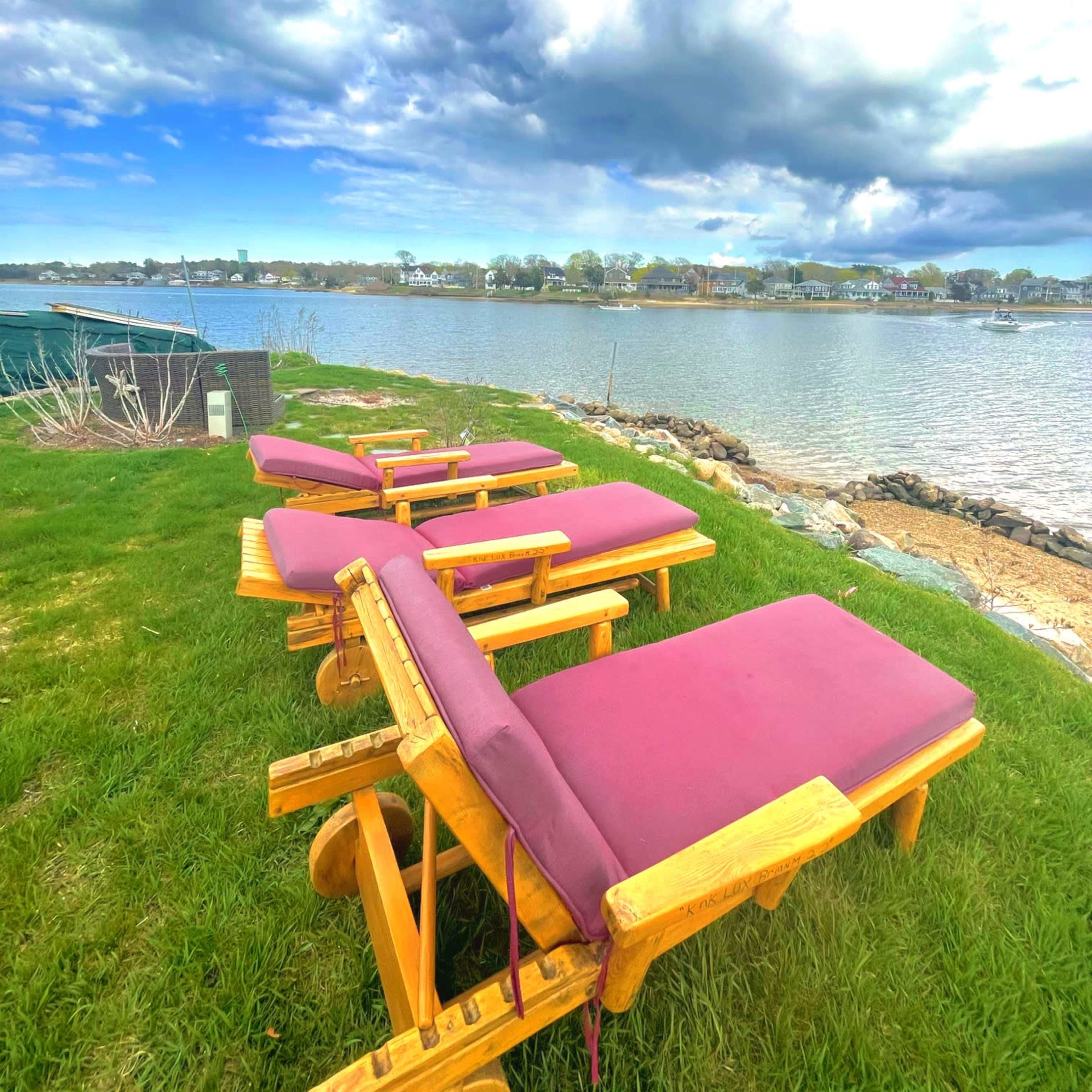 Four wooden lounge chairs with purple cushions are lined up on a grassy area beside a calm waterway under a partly cloudy sky.