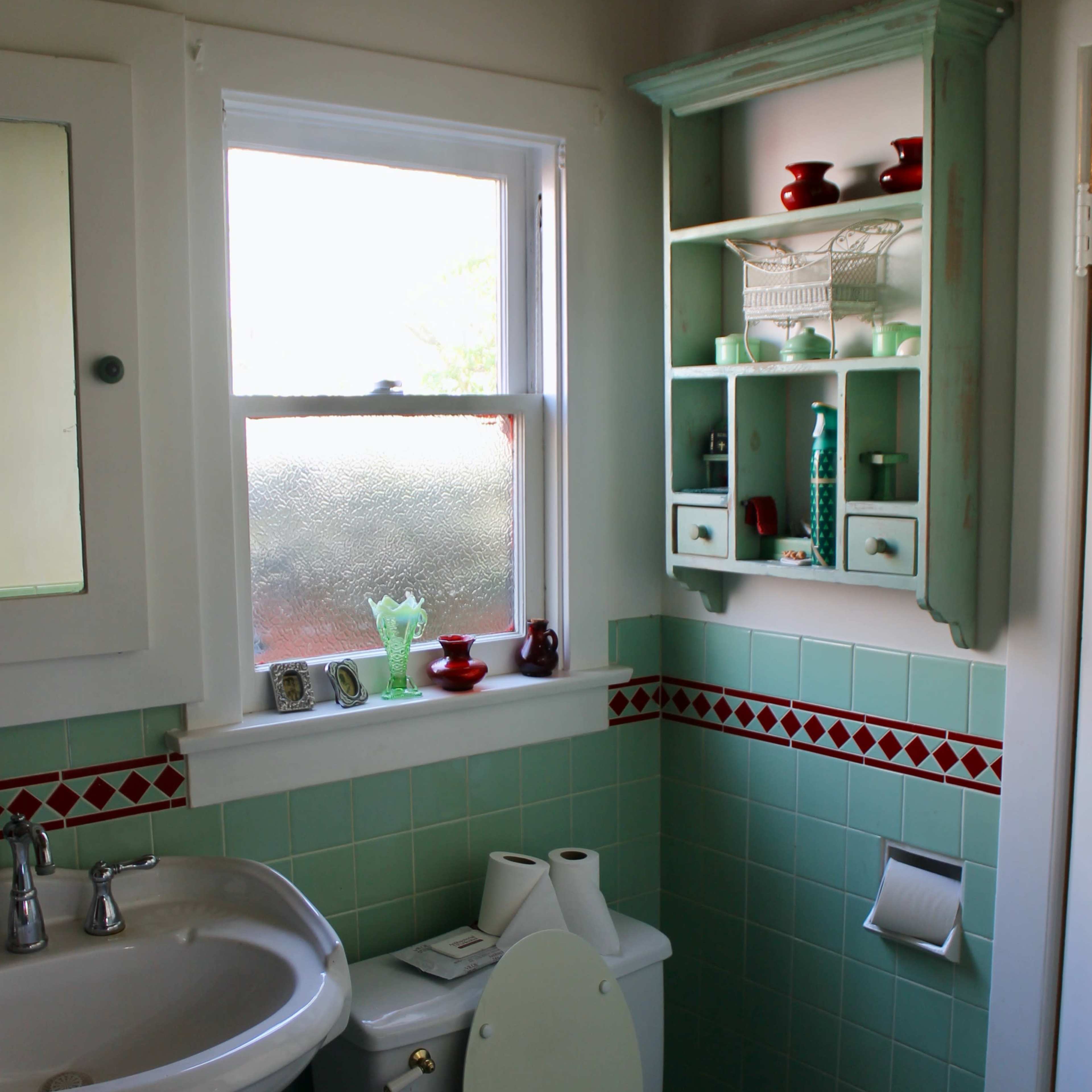 A small bathroom features a white pedestal sink, a toilet, and a wall-mounted cabinet with various decorative items, set against teal tile and a patterned border.