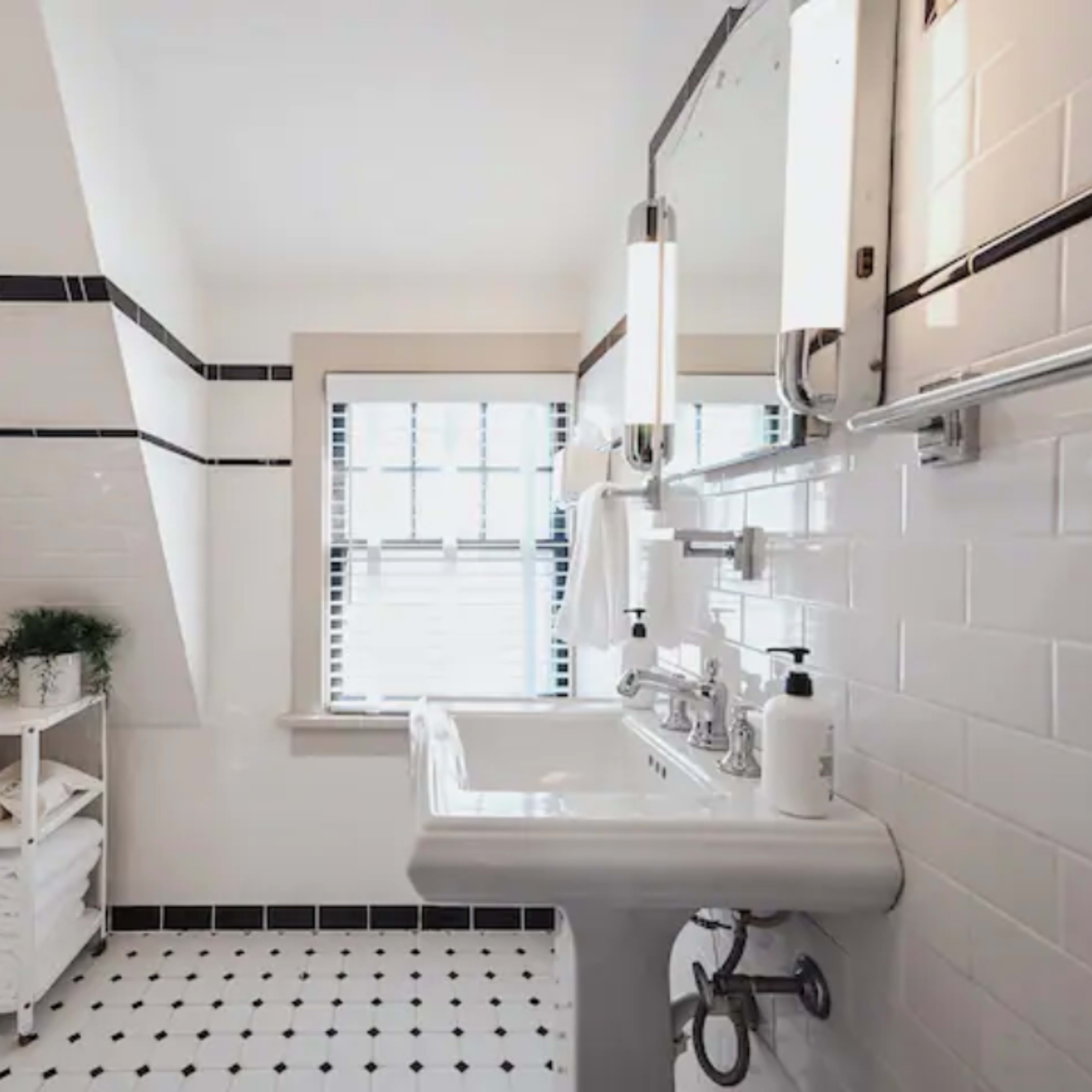 The bathroom features a white tiled wall with black accents, a pedestal sink, and a window with horizontally striped blinds.