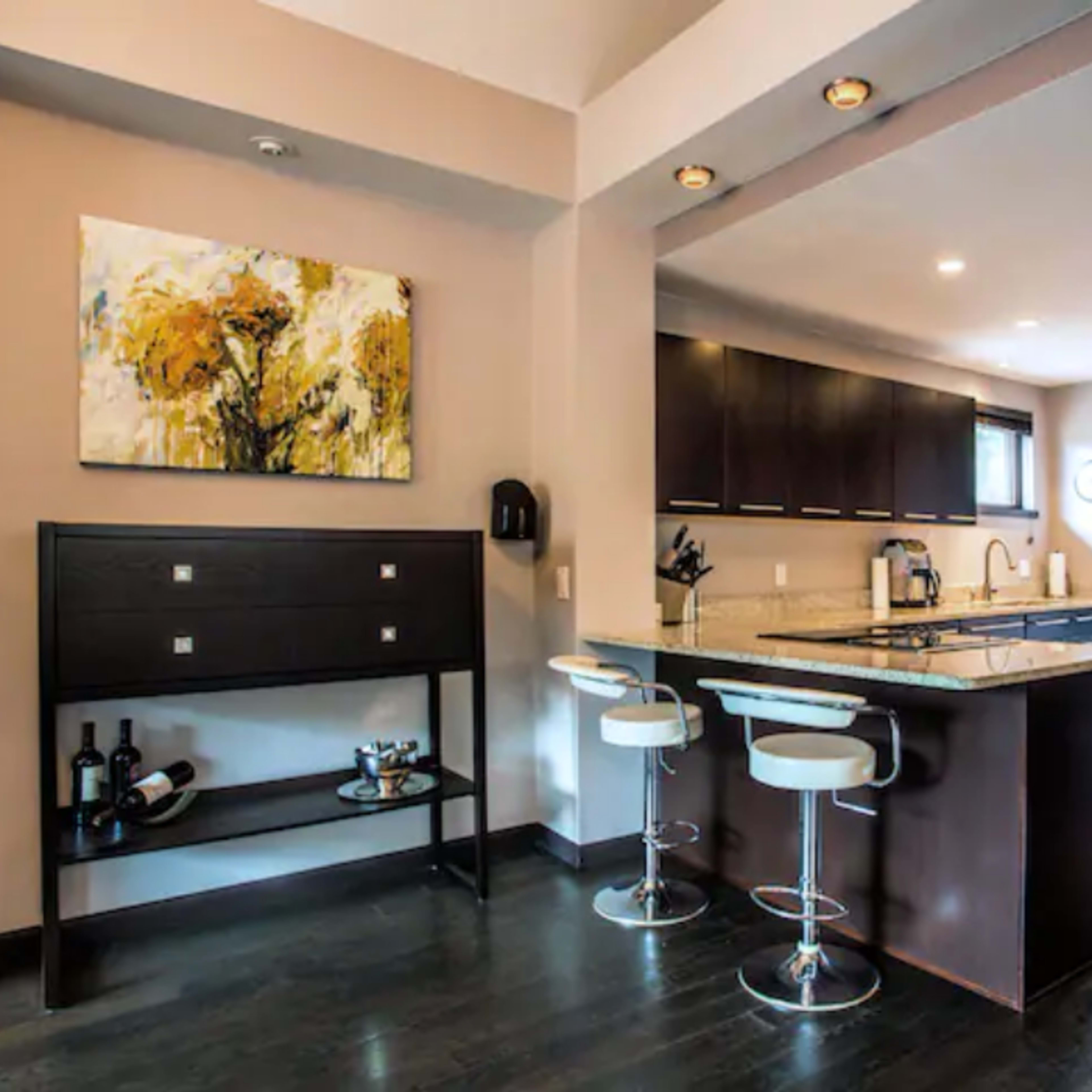 A modern kitchen with dark wood cabinets, a granite countertop, and two white bar stools, alongside a dining area with an abstract painting and a small console table.