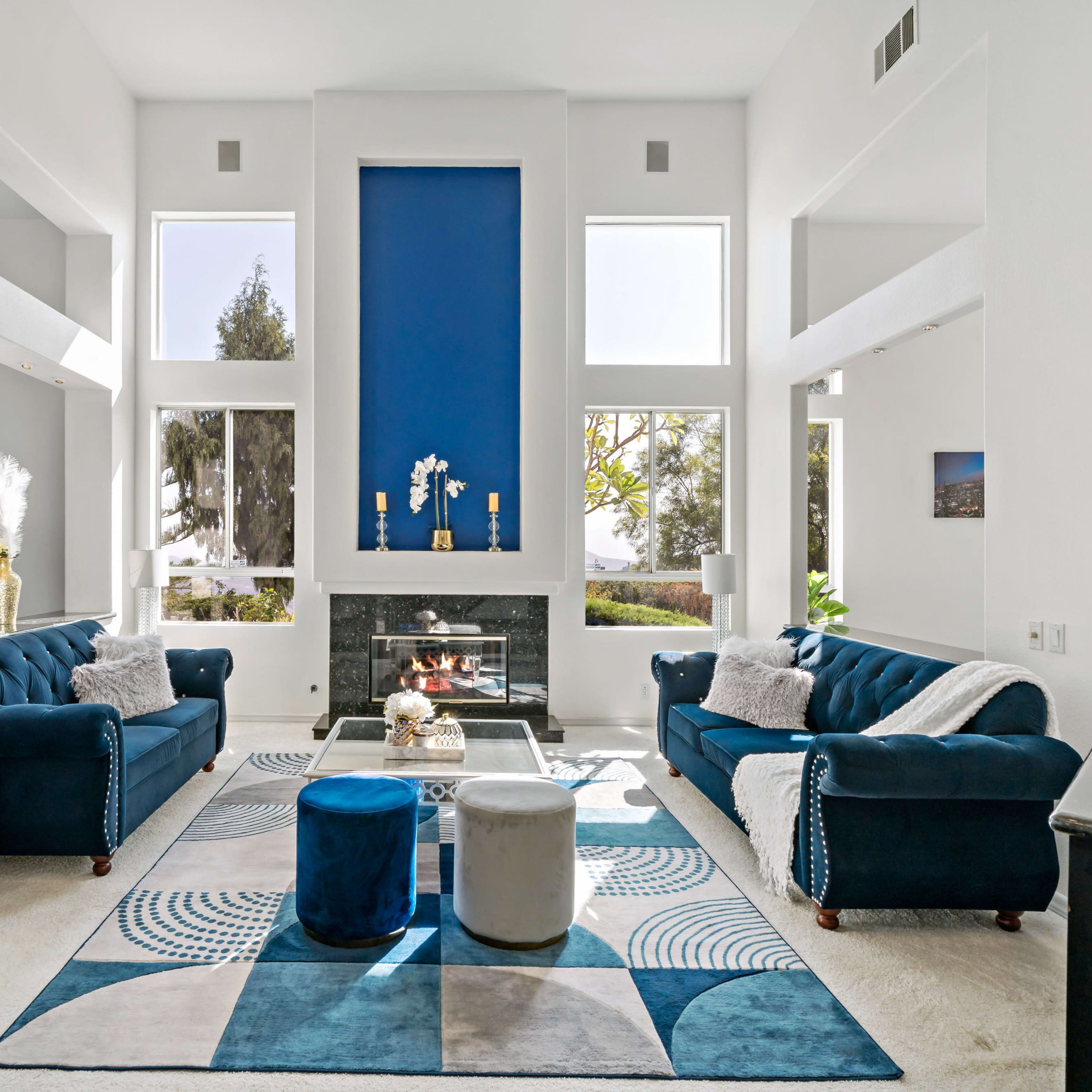 The living room features two blue sofas facing a fireplace, with large windows allowing natural light to fill the space and a geometric rug on the floor.
