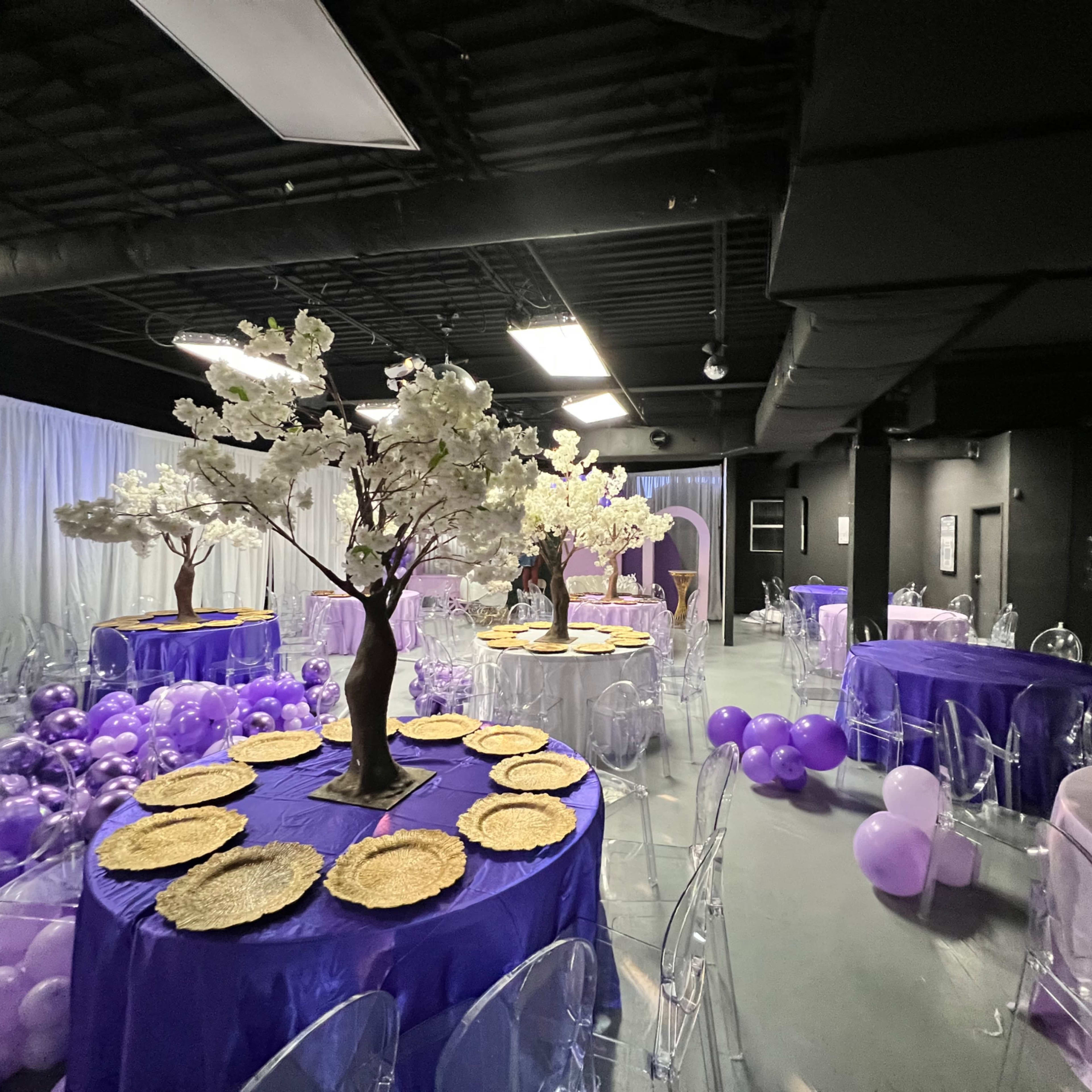 An event space with round tables covered in purple tablecloths, decorated with floral centerpieces and surrounded by clusters of purple balloons.