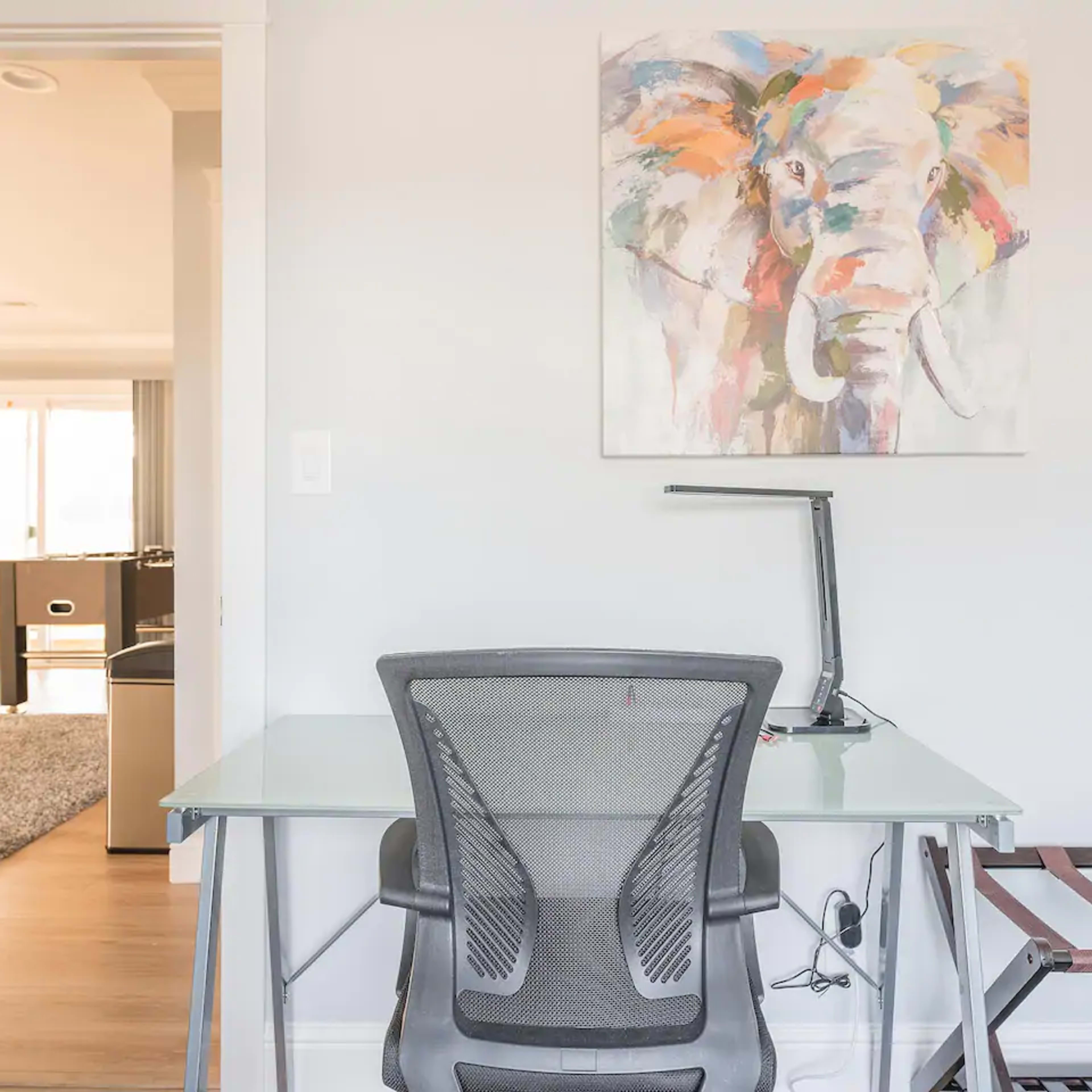 A modern workspace features a glass desk with a mesh chair, illuminated by a lamp, beside a wall adorned with a colorful elephant painting.