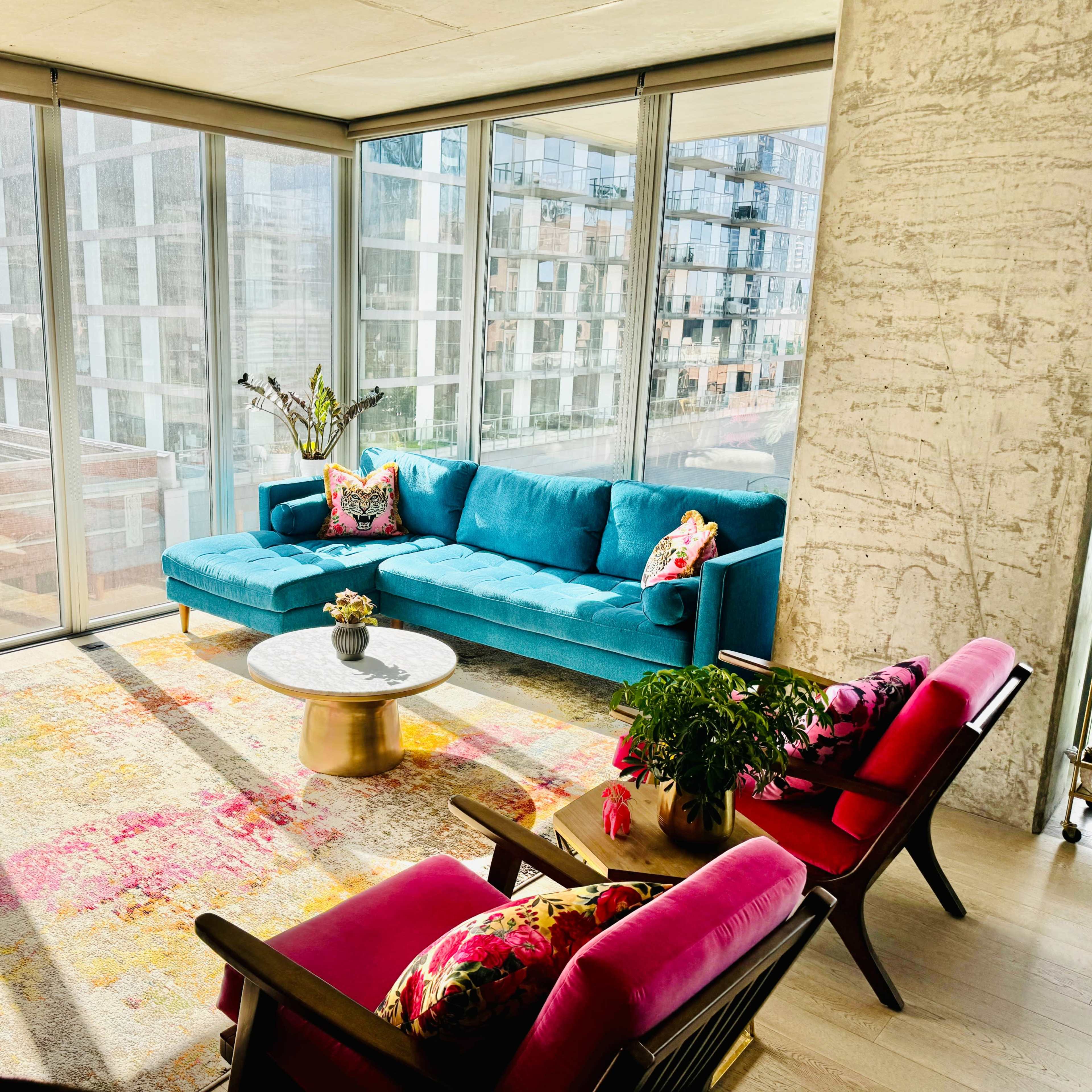 A bright living room features a blue sofa, colorful chairs, a round coffee table, and large windows overlooking a cityscape.