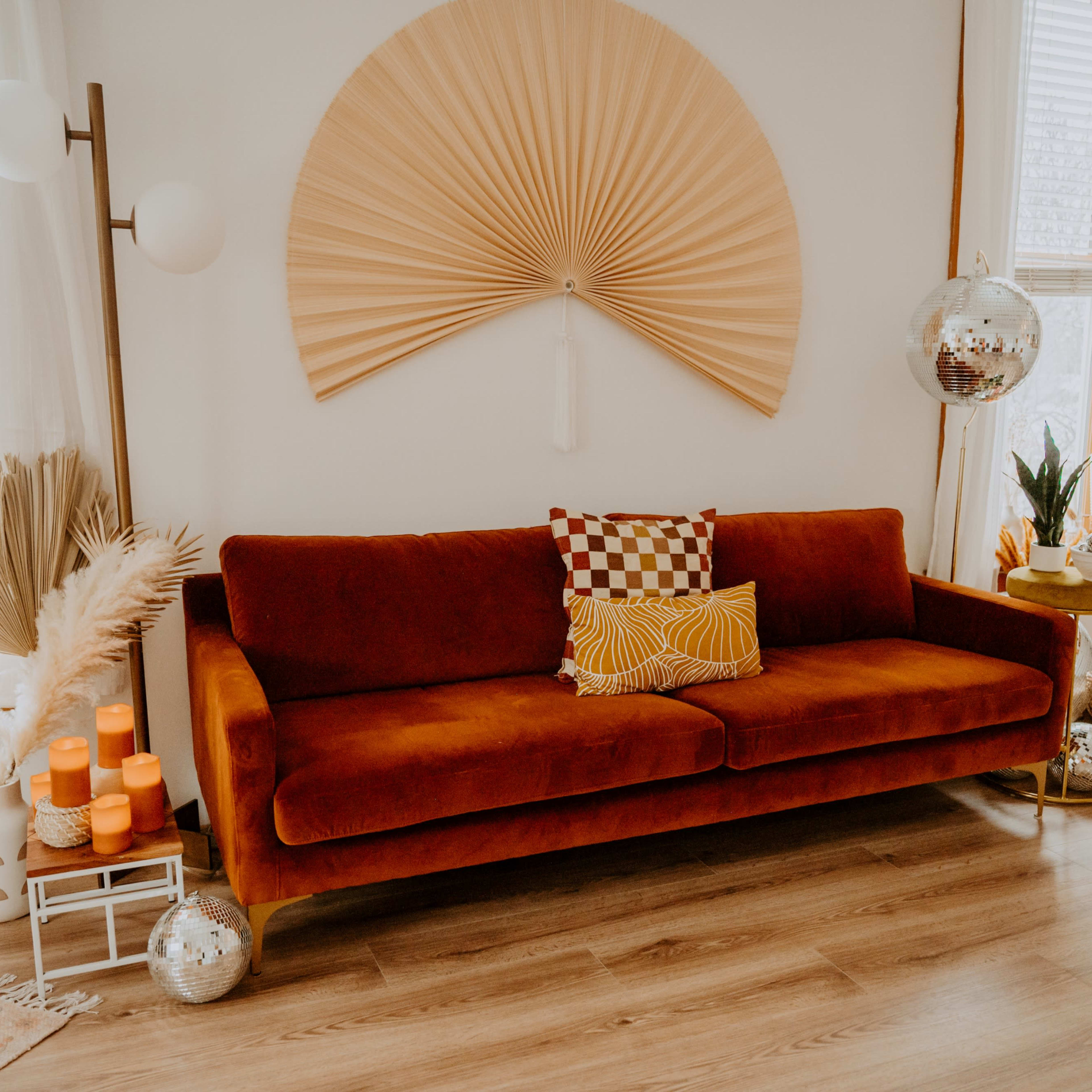 A cozy living room with an orange velvet sofa, decorative wall art, potted plants, and various accessories arranged on the floor and nearby surfaces.