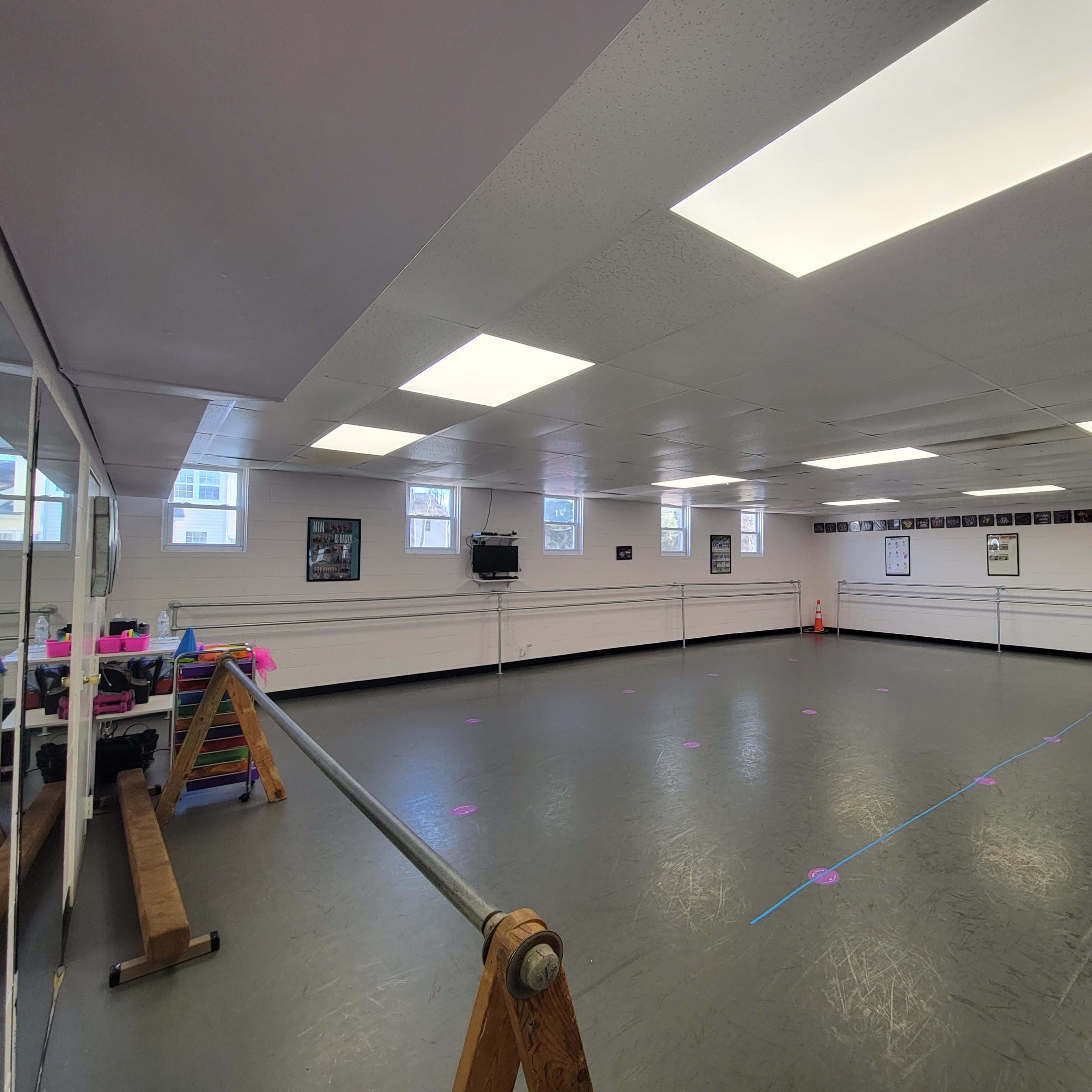 An empty dance studio with a mirrored wall, wooden ballet barre, and bright overhead lights.