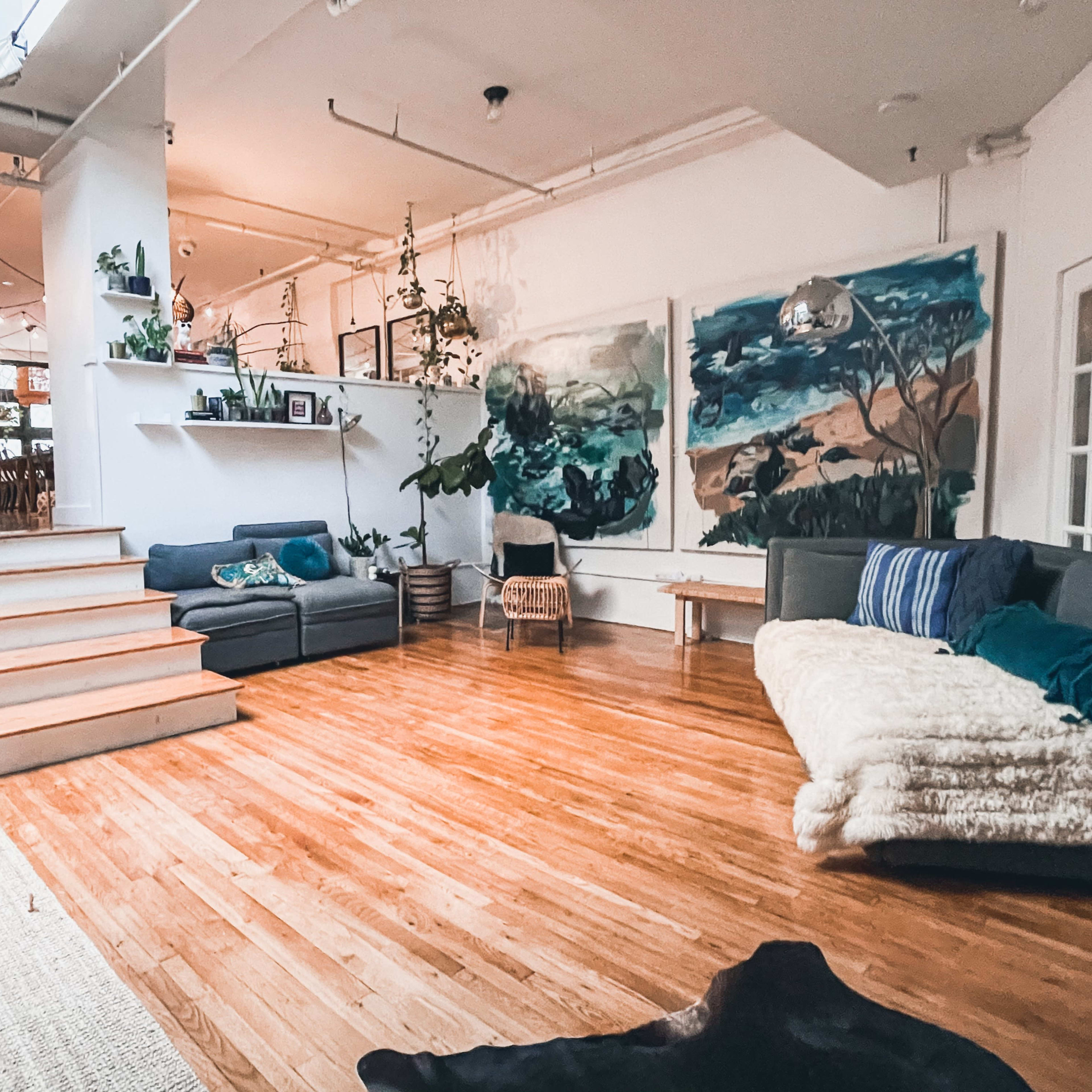 A spacious, well-lit room featuring wooden floors, a stairway leading to a higher level, and several plants arranged throughout the space, with large wall paintings in the background.