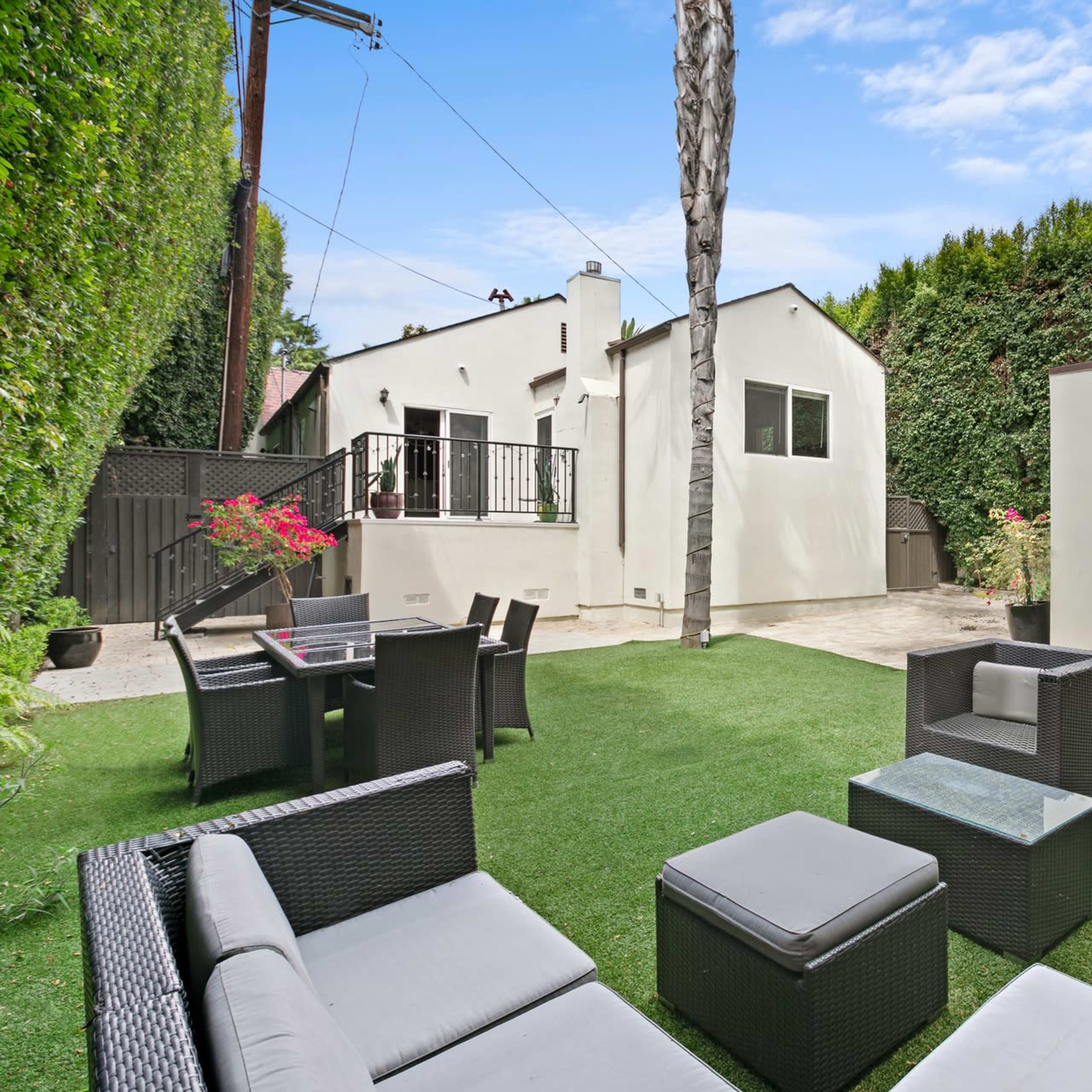 A landscaped backyard with modern outdoor seating, a small dining area, and two white buildings in the background.