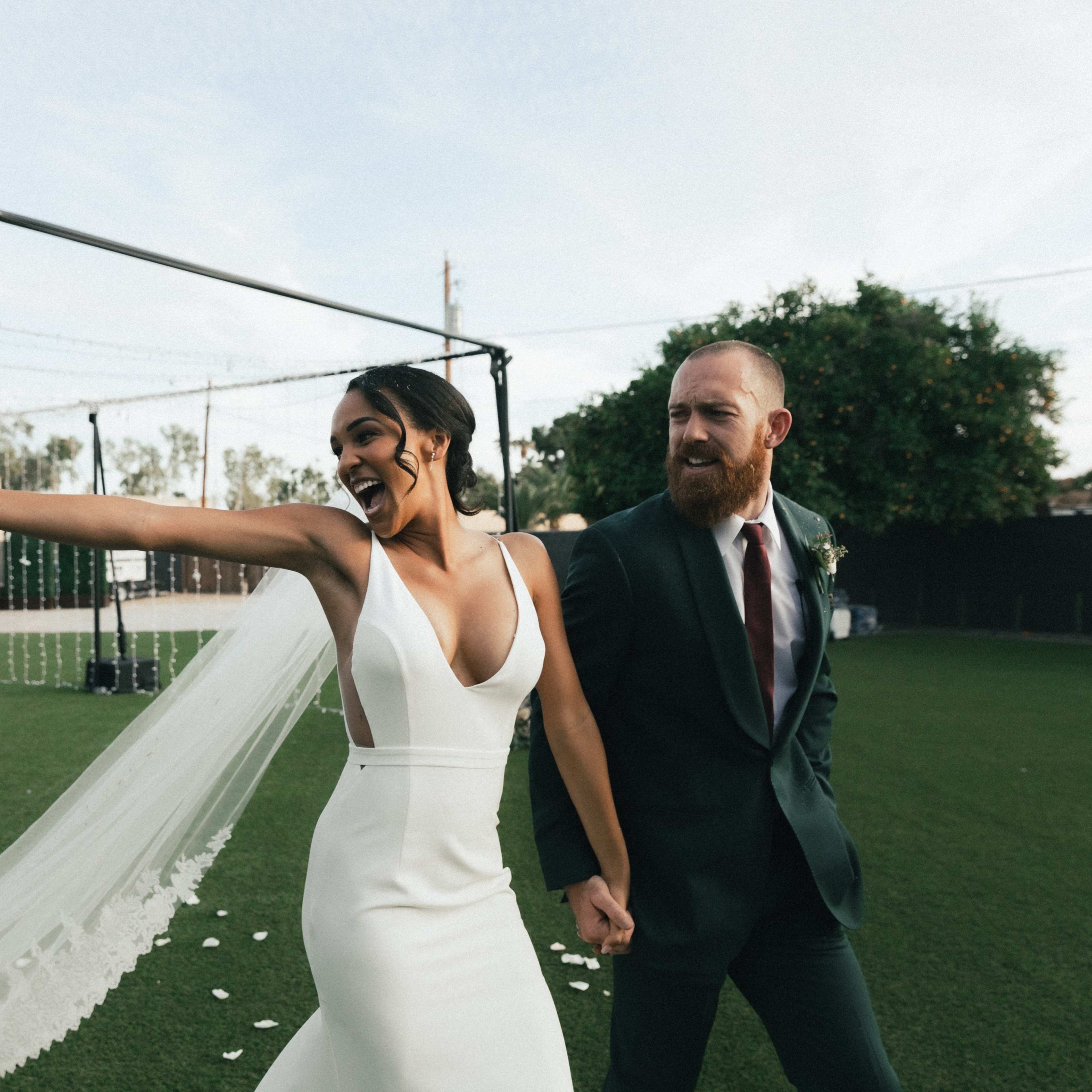 A couple celebrates their wedding by holding hands and smiling as they walk together in an outdoor venue.