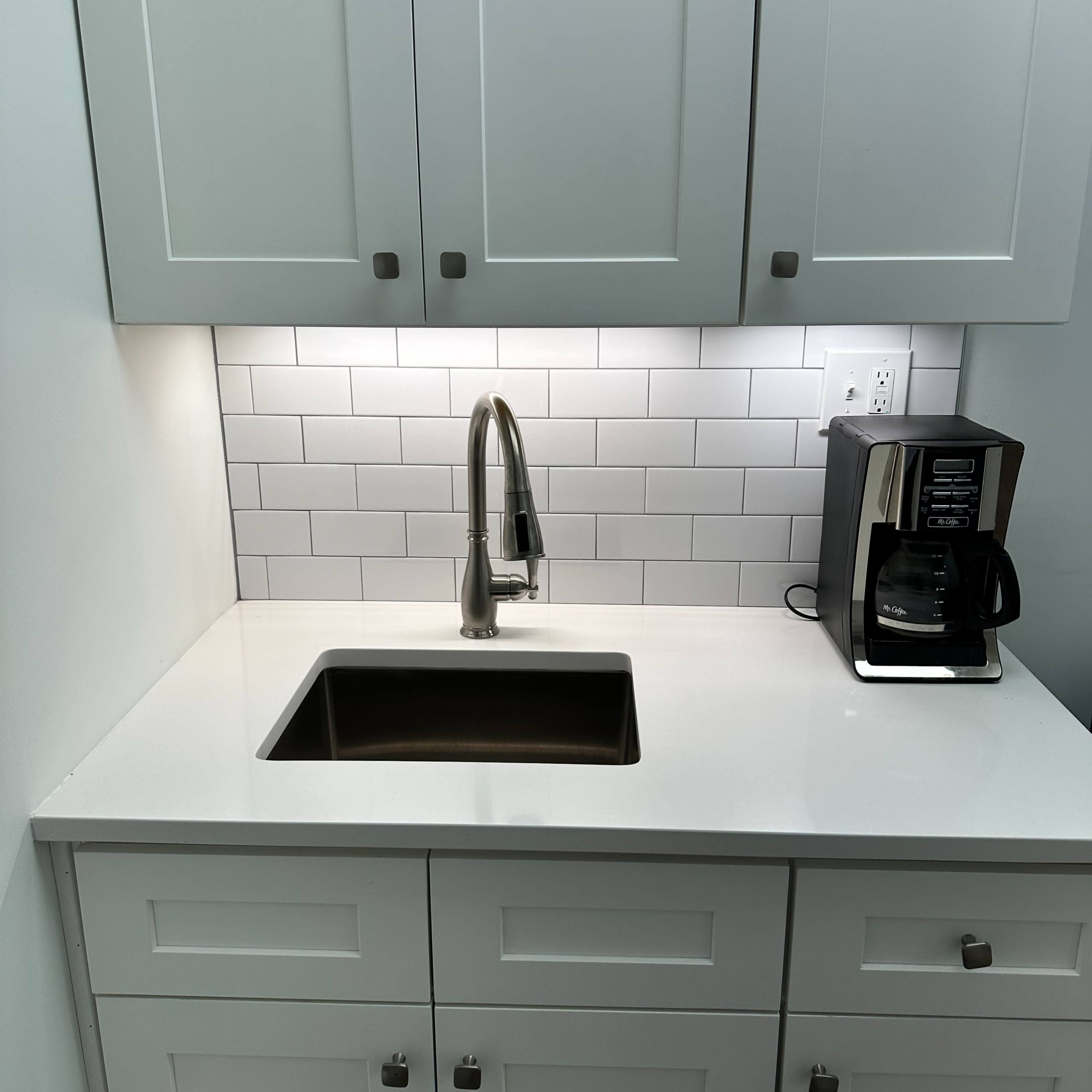 A modern kitchenette with white cabinets, a stainless steel sink, and a coffee maker on the counter.