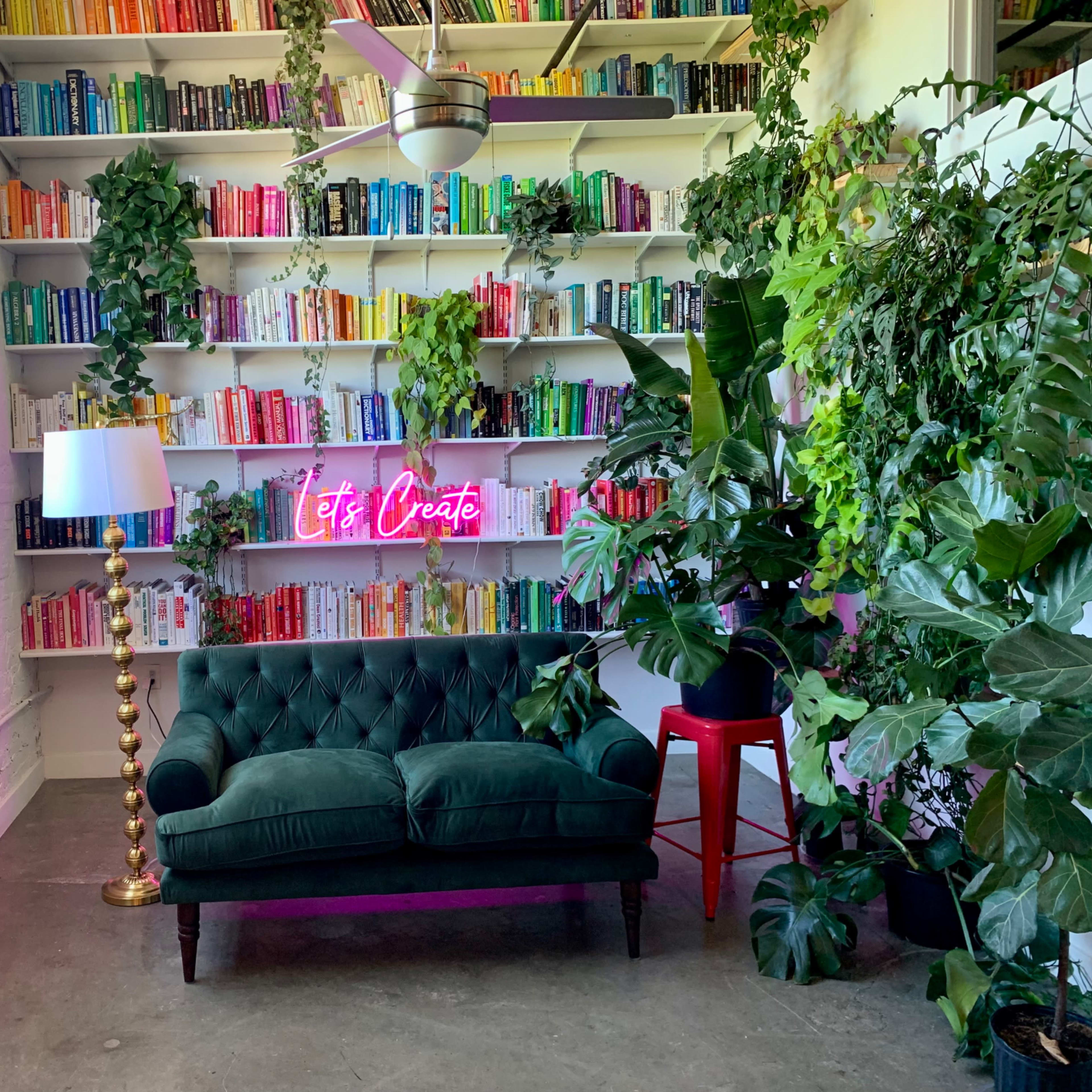 A green tufted sofa sits in front of a brightly colored bookshelf, surrounded by various plants and illuminated by a neon sign that reads "Let's Create."