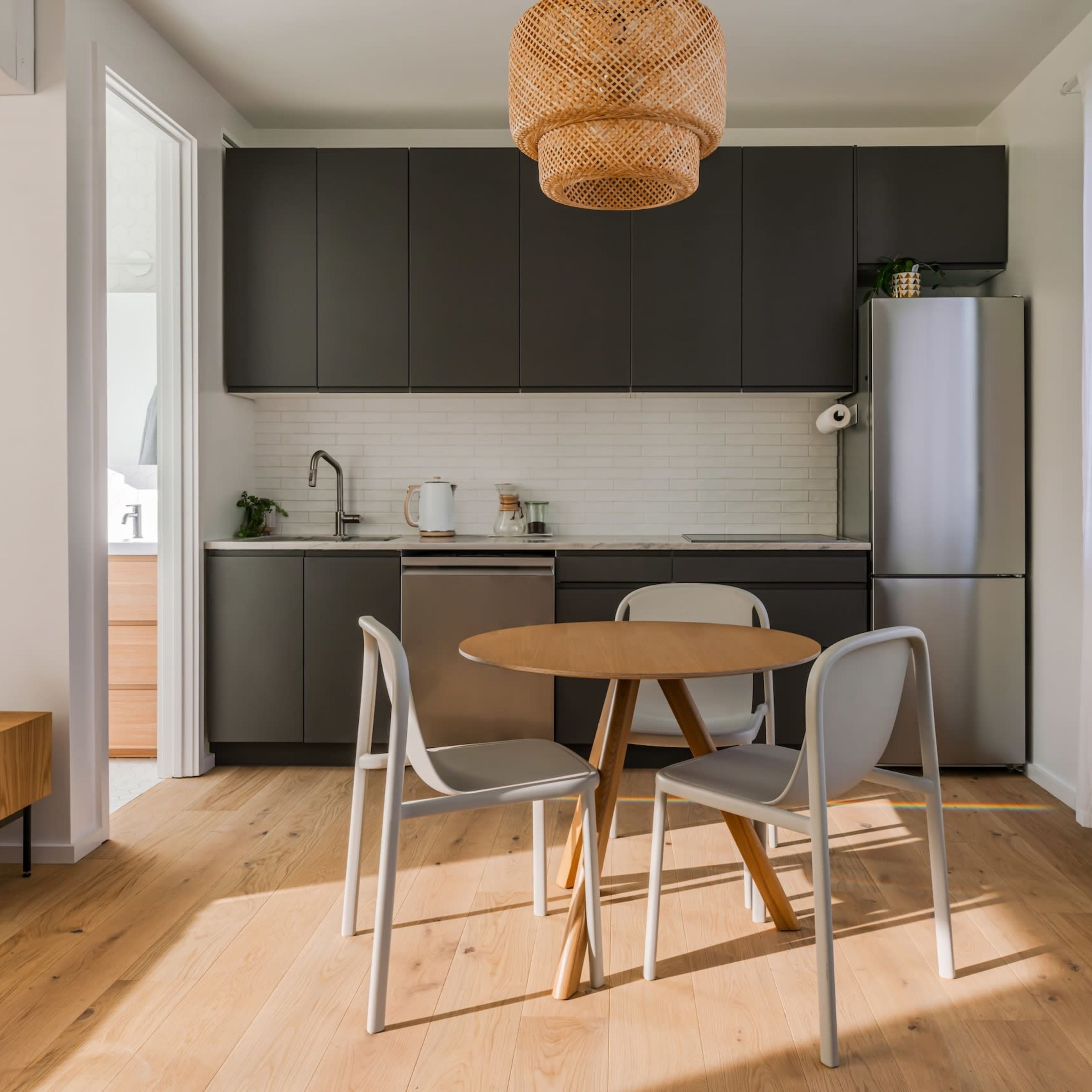 A modern kitchen features dark cabinetry, a stainless steel refrigerator, a round dining table with three chairs, and natural light streaming through sheer curtains.