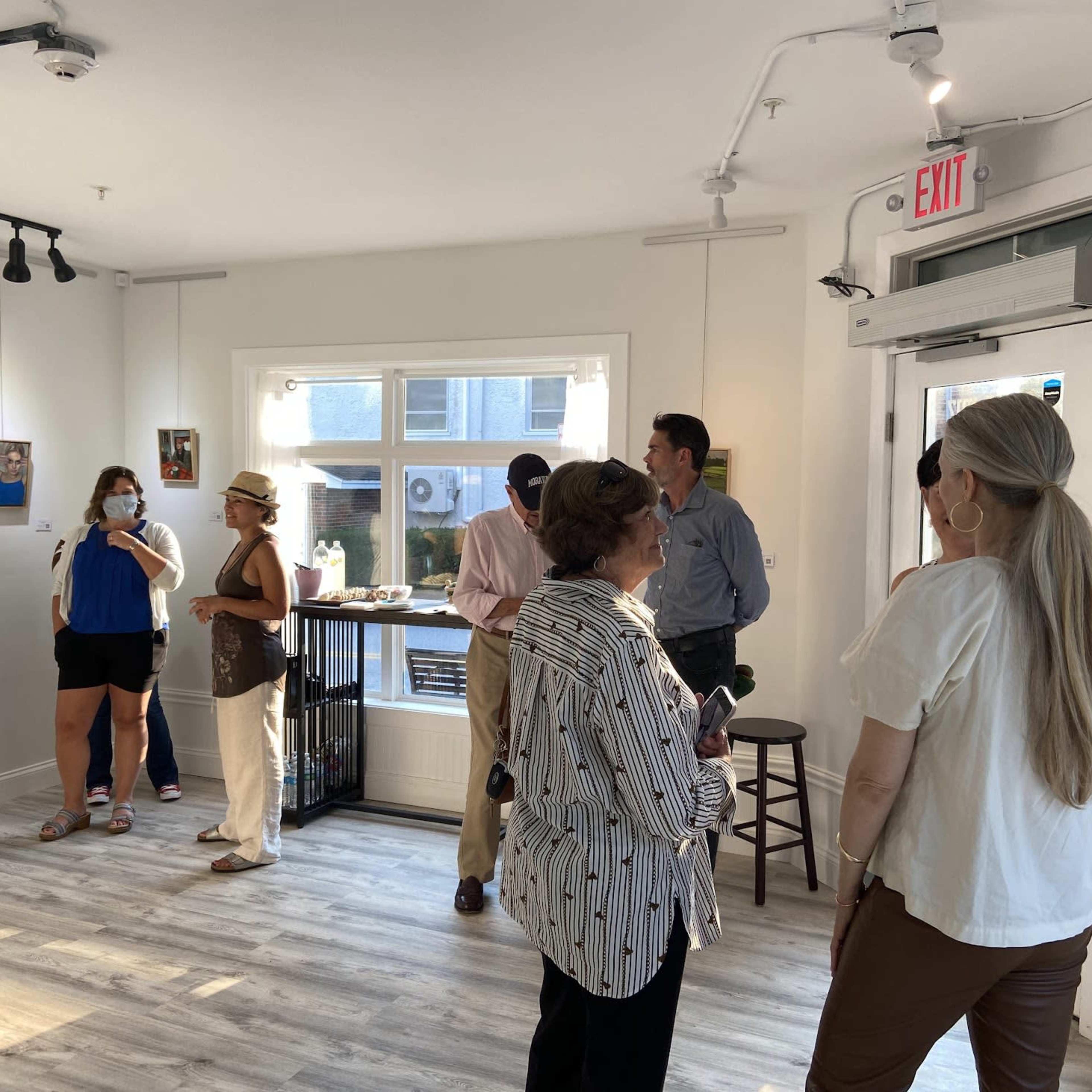 A group of people is gathered in a brightly lit art gallery, with several artworks displayed on the walls.