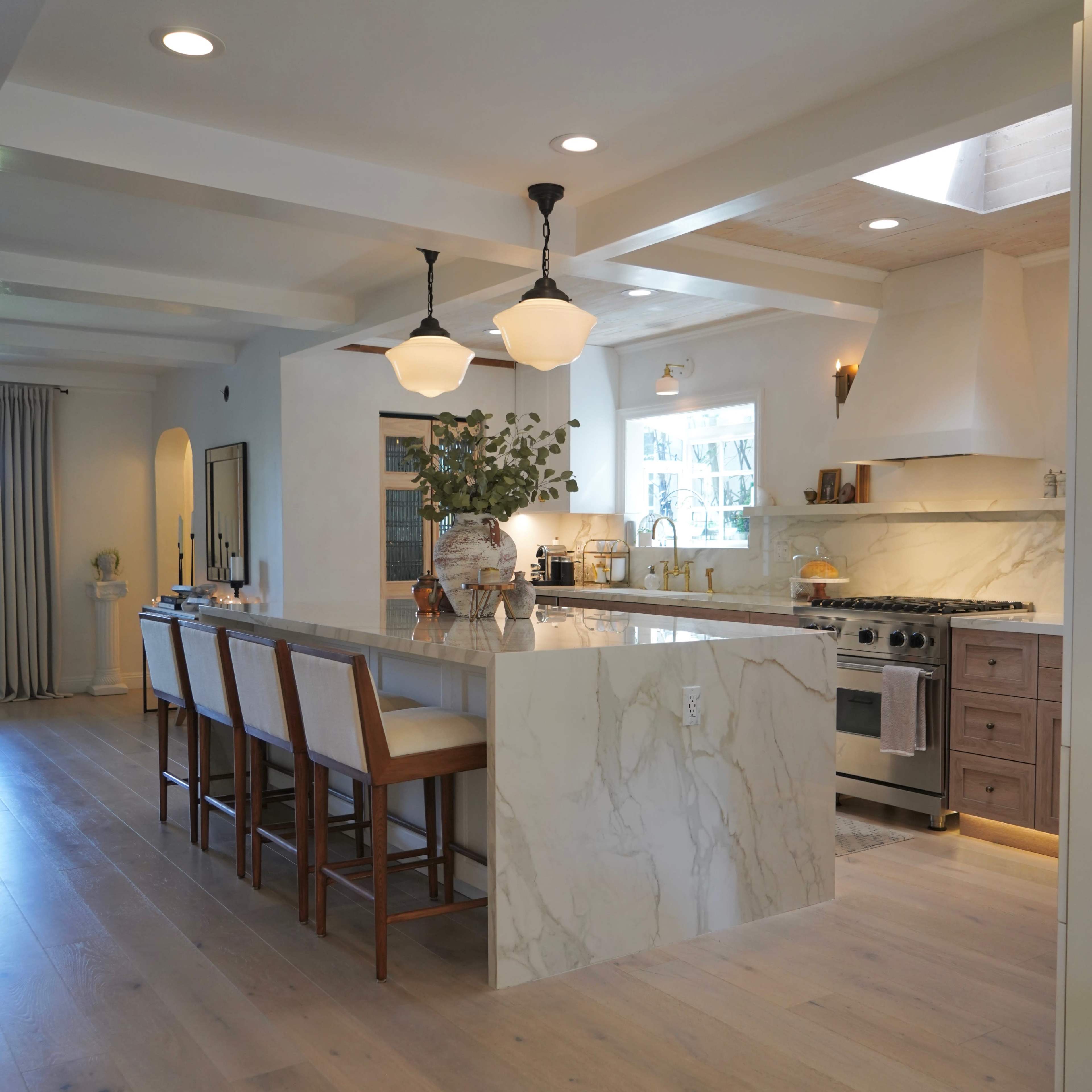A modern kitchen with a spacious island featuring marble countertops, pendant lighting, and a stove with an exhaust hood, surrounded by wooden barstools.
