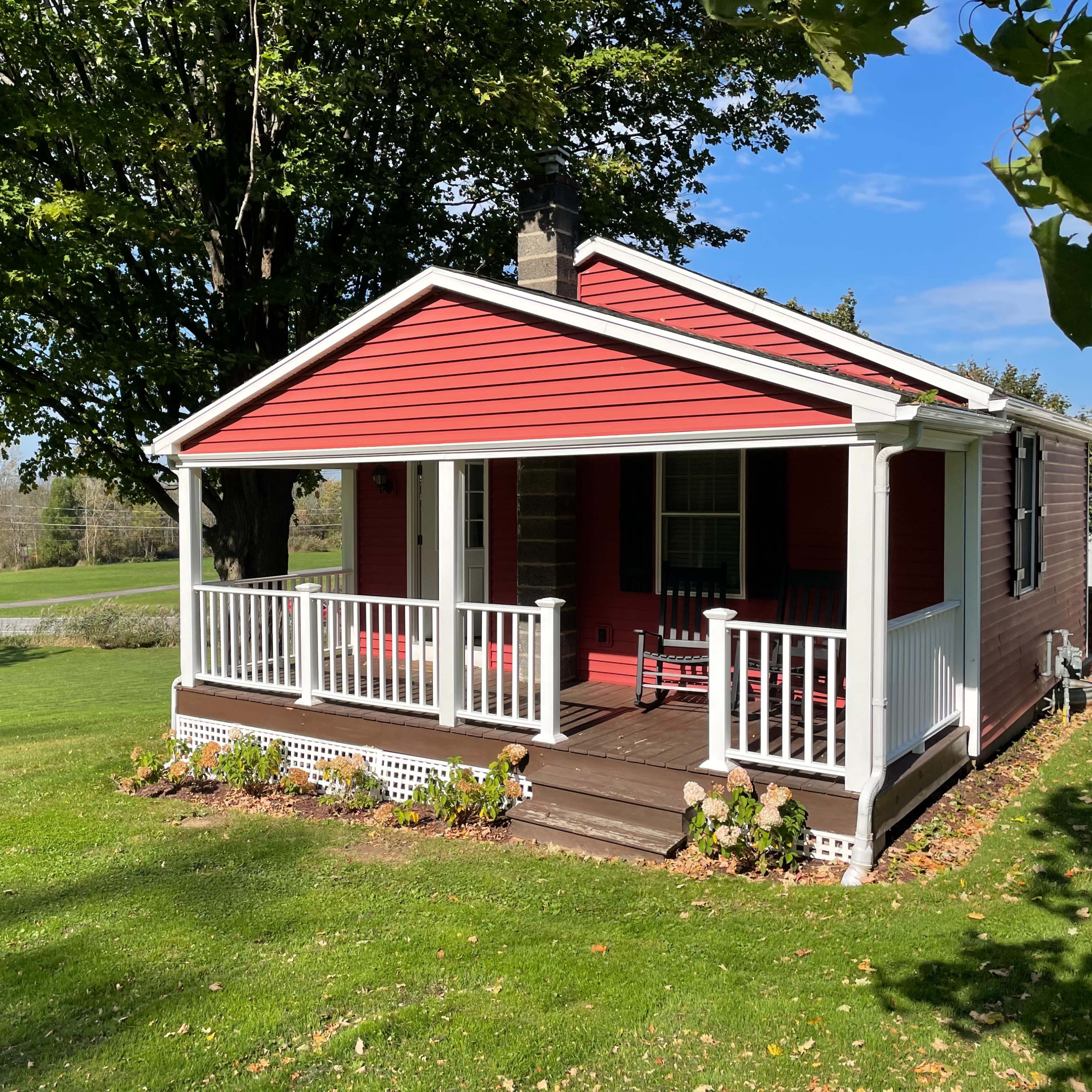 A small red house with a white porch and railing sits on a grassy lawn surrounded by trees.