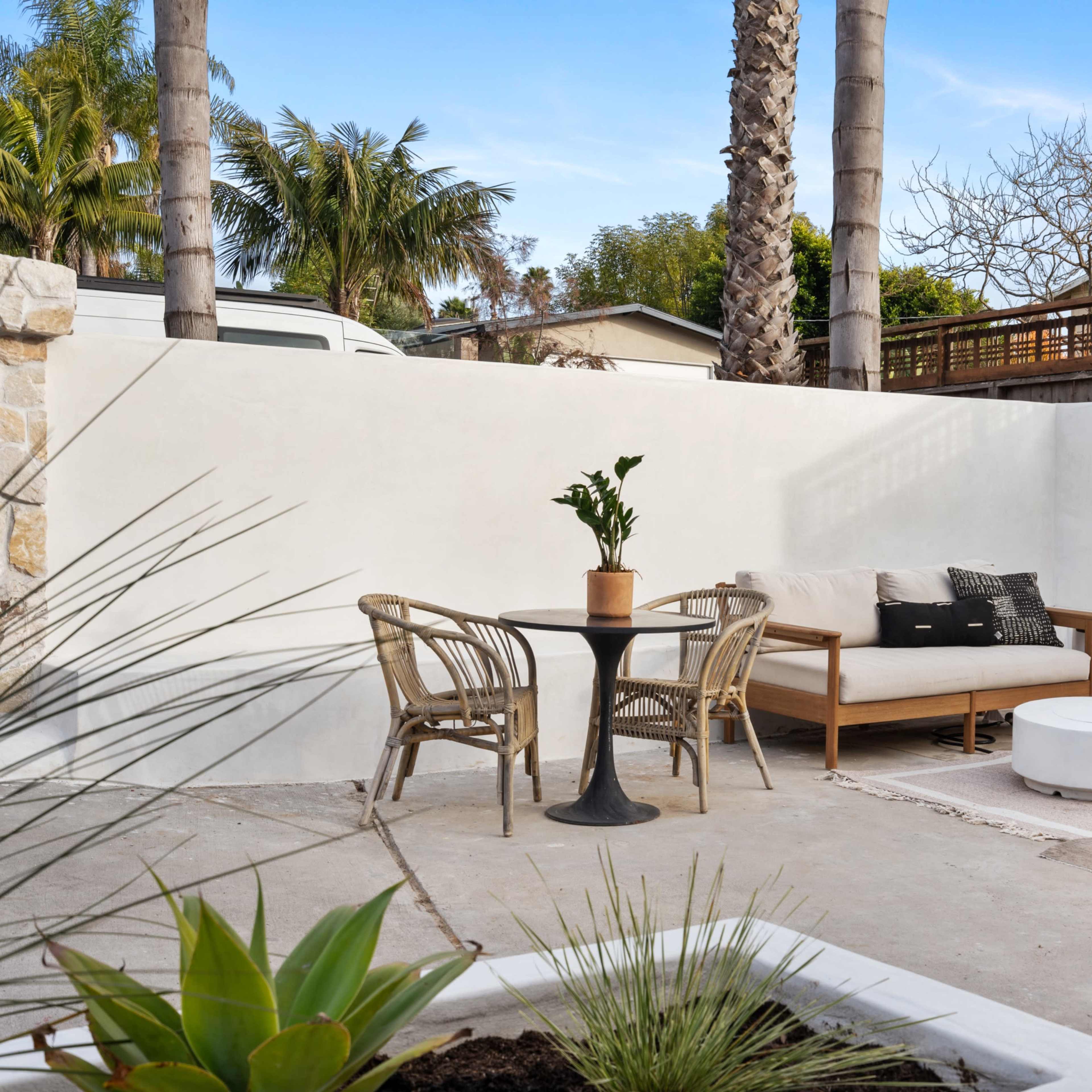 A minimalist outdoor seating area with a wooden couch, a round table, and potted plants, surrounded by white walls and palm trees.