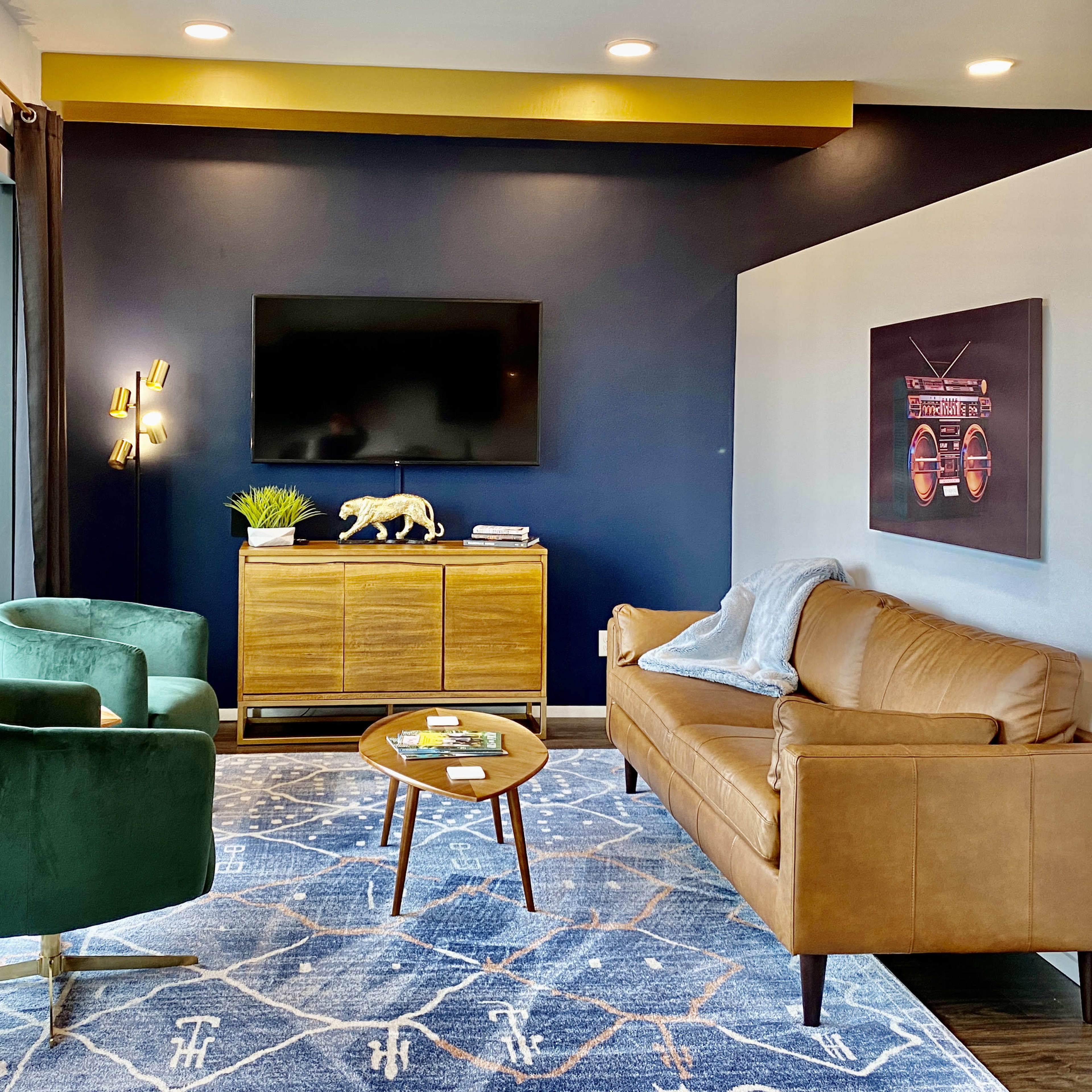 The living room features a leather sofa, two green armchairs, a wooden sideboard, a television mounted on the wall, and a patterned area rug.